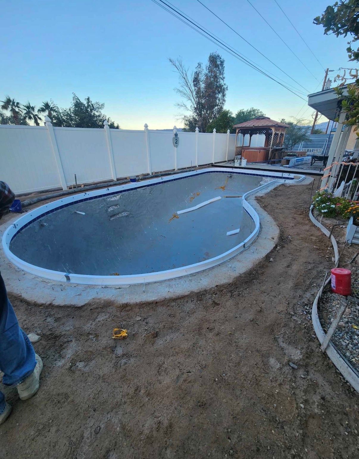 Pool under construction with concrete border, tan dirt, and white fence.