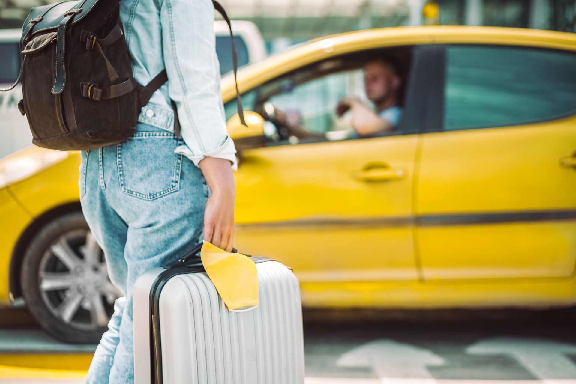 A woman is walking with a suitcase and a backpack in front of a yellow taxi.