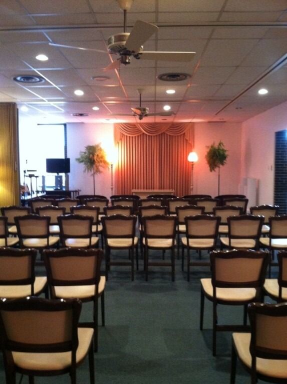 Empty chapel with rows of chairs, stage with curtain, ceiling lights, and two small trees.