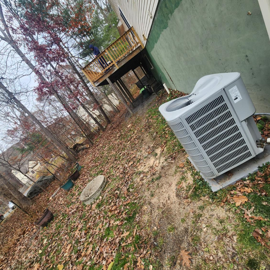 An outdoor HVAC unit sits on a concrete pad next to the side of a house near a wooden deck and a wooded backyard.