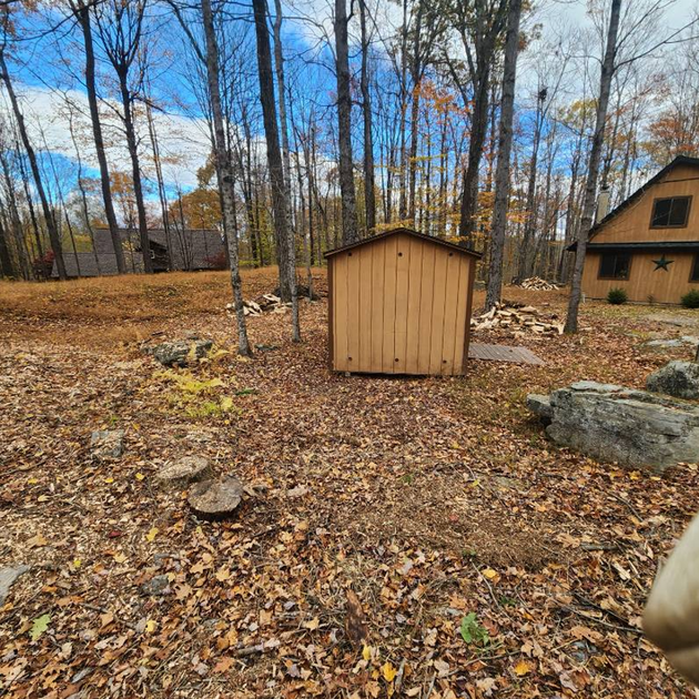 A tan wooden shed sits on a leaf-covered forest floor, with a larger house visible in the background among trees.