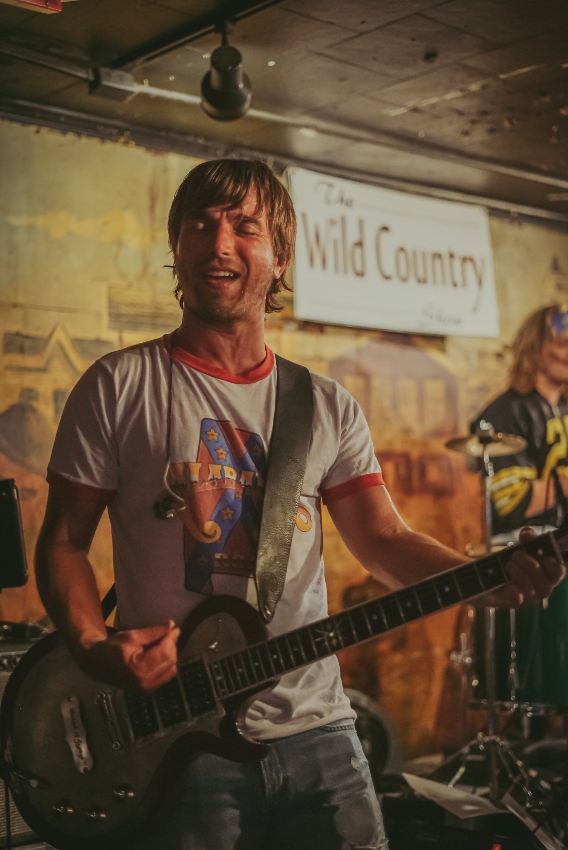 A man is playing a guitar in front of a sign that says wild country.