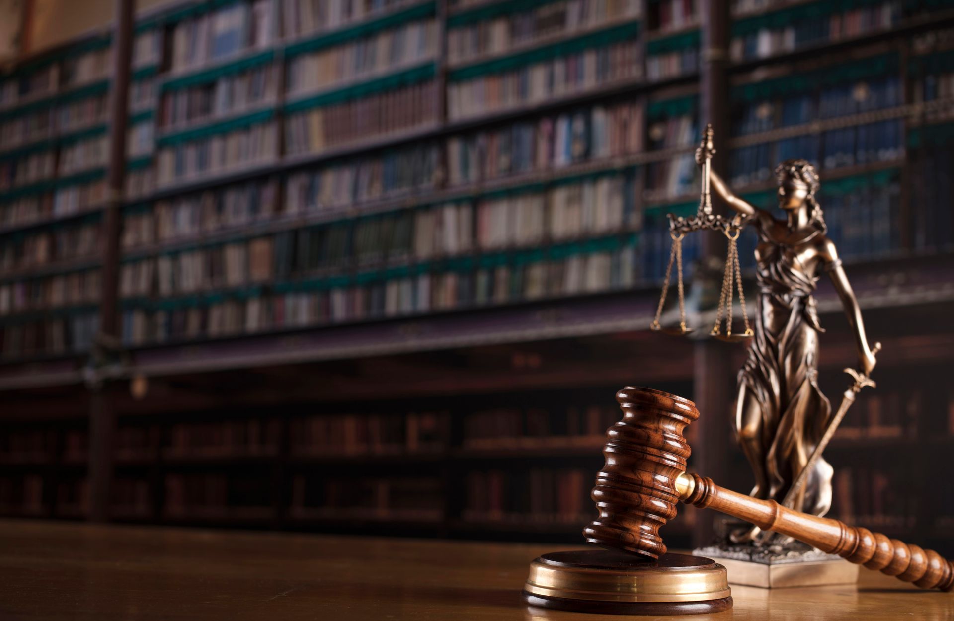 Gavel next to Lady Justice statue with scales and sword, in a law library with bookshelves.