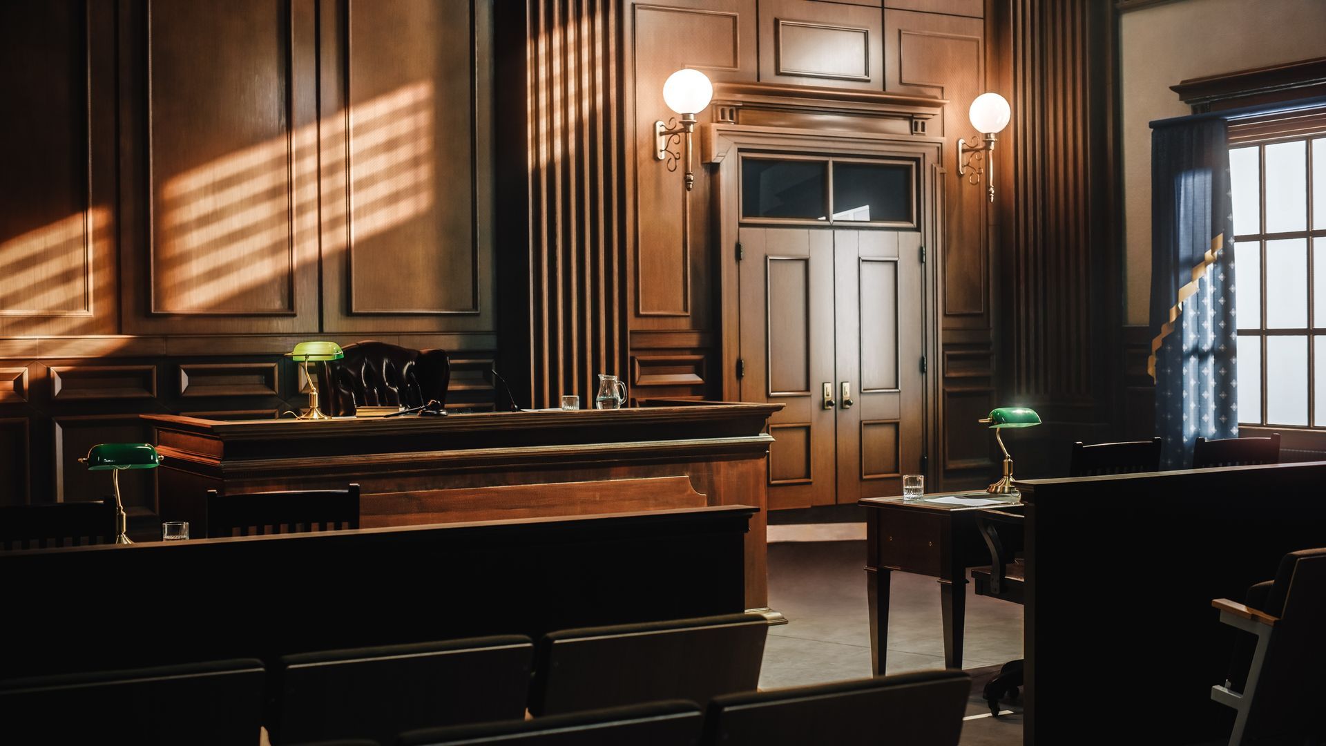 Empty courtroom with wooden paneling, a judge's bench, and rows of seats, illuminated by sunlight.