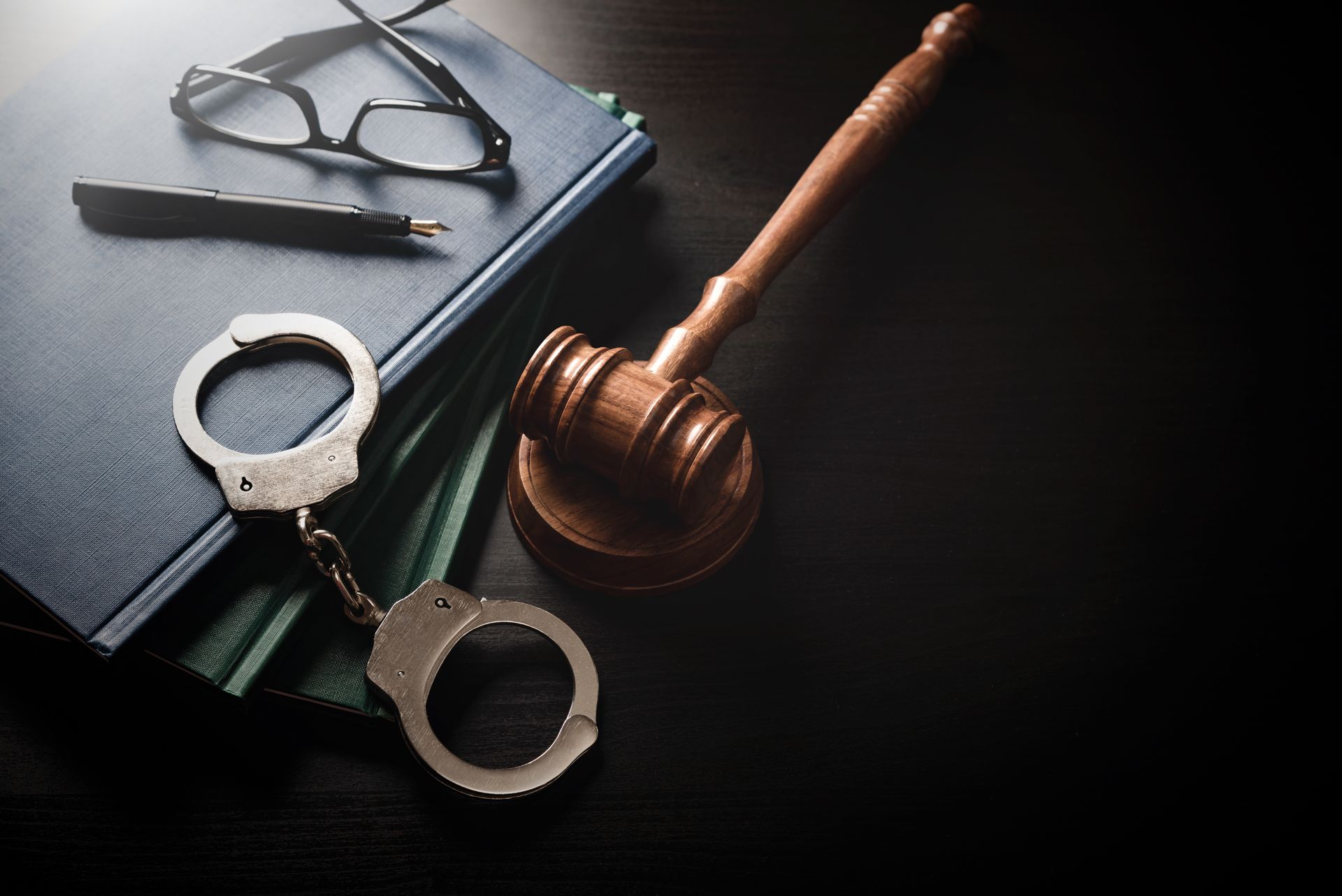 Handcuffs, gavel, books, glasses, and pen on a dark surface, representing legal or judicial context.