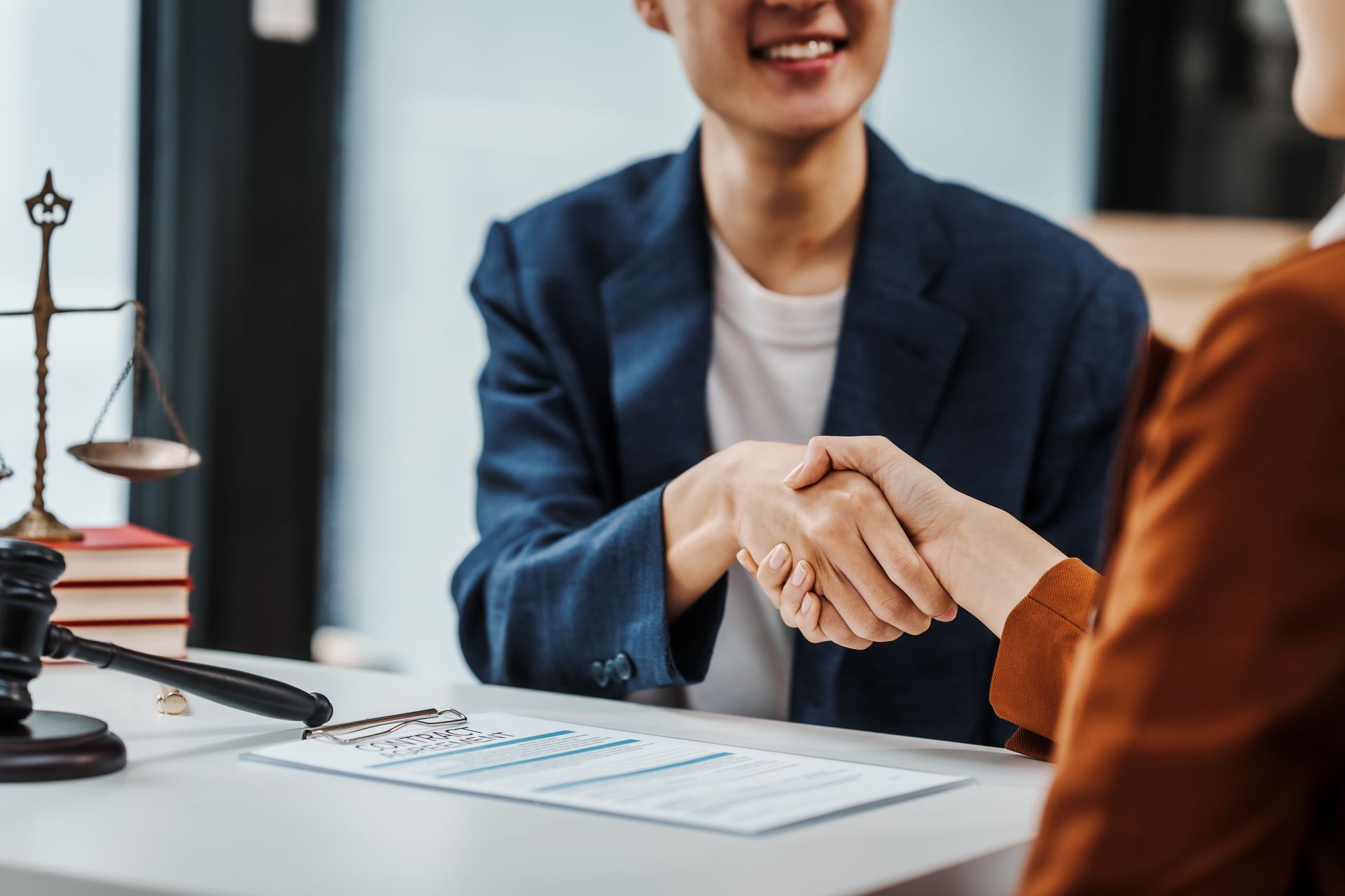 Two people shaking hands over a document at a desk with law scales and a gavel. One smiles.
