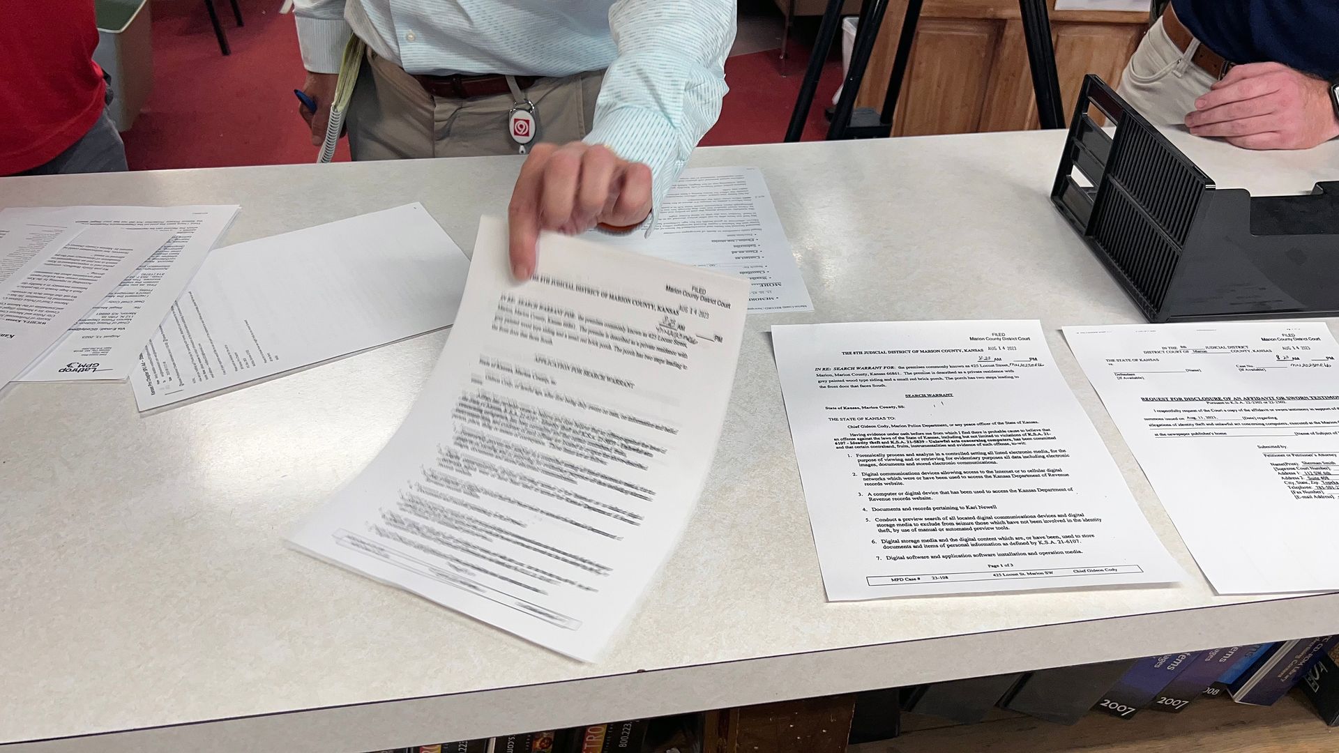 Person pointing at a document on a table with other papers, a printer, and another person visible.