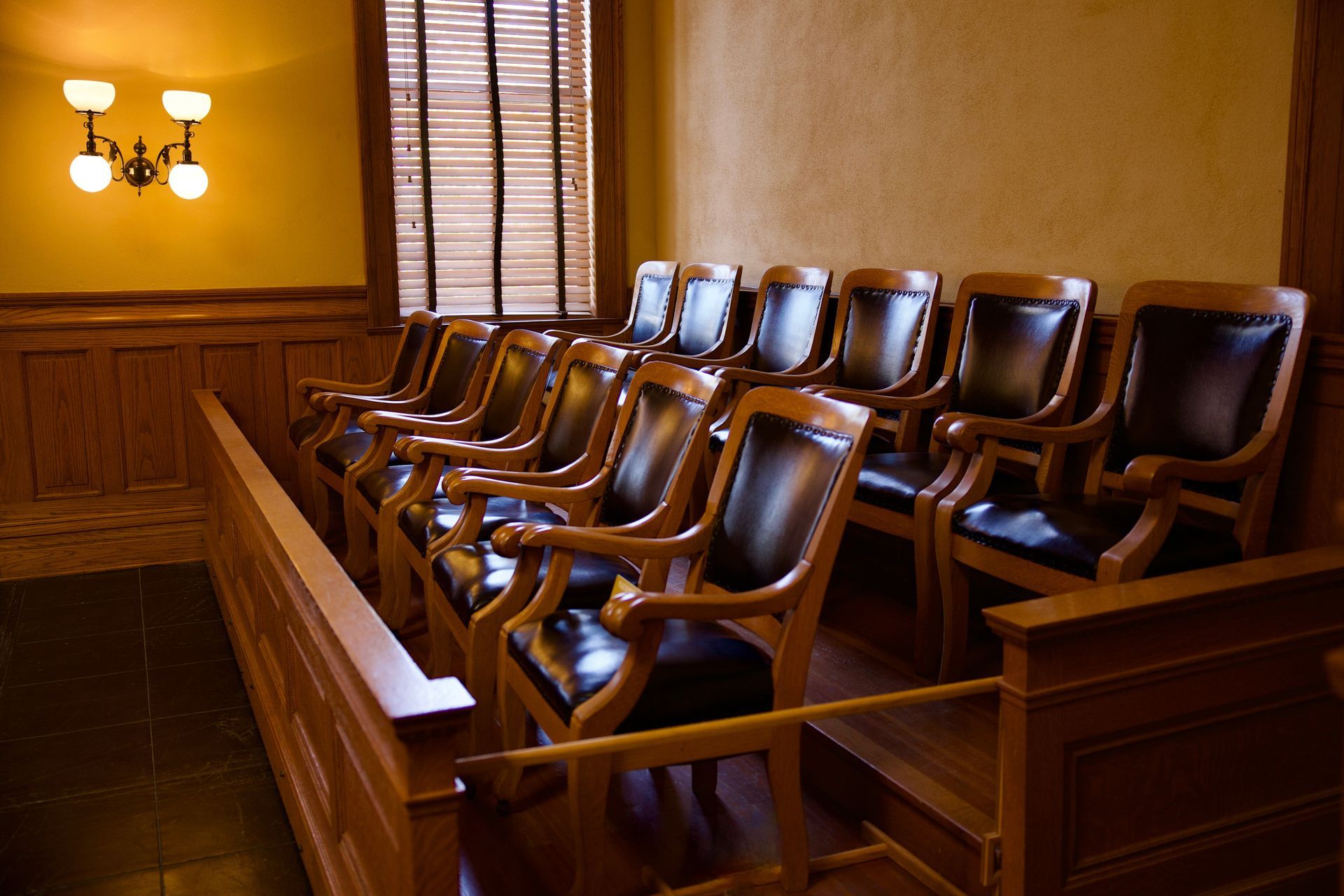 Empty jury box in a courtroom with wooden chairs, railing, and blinds.