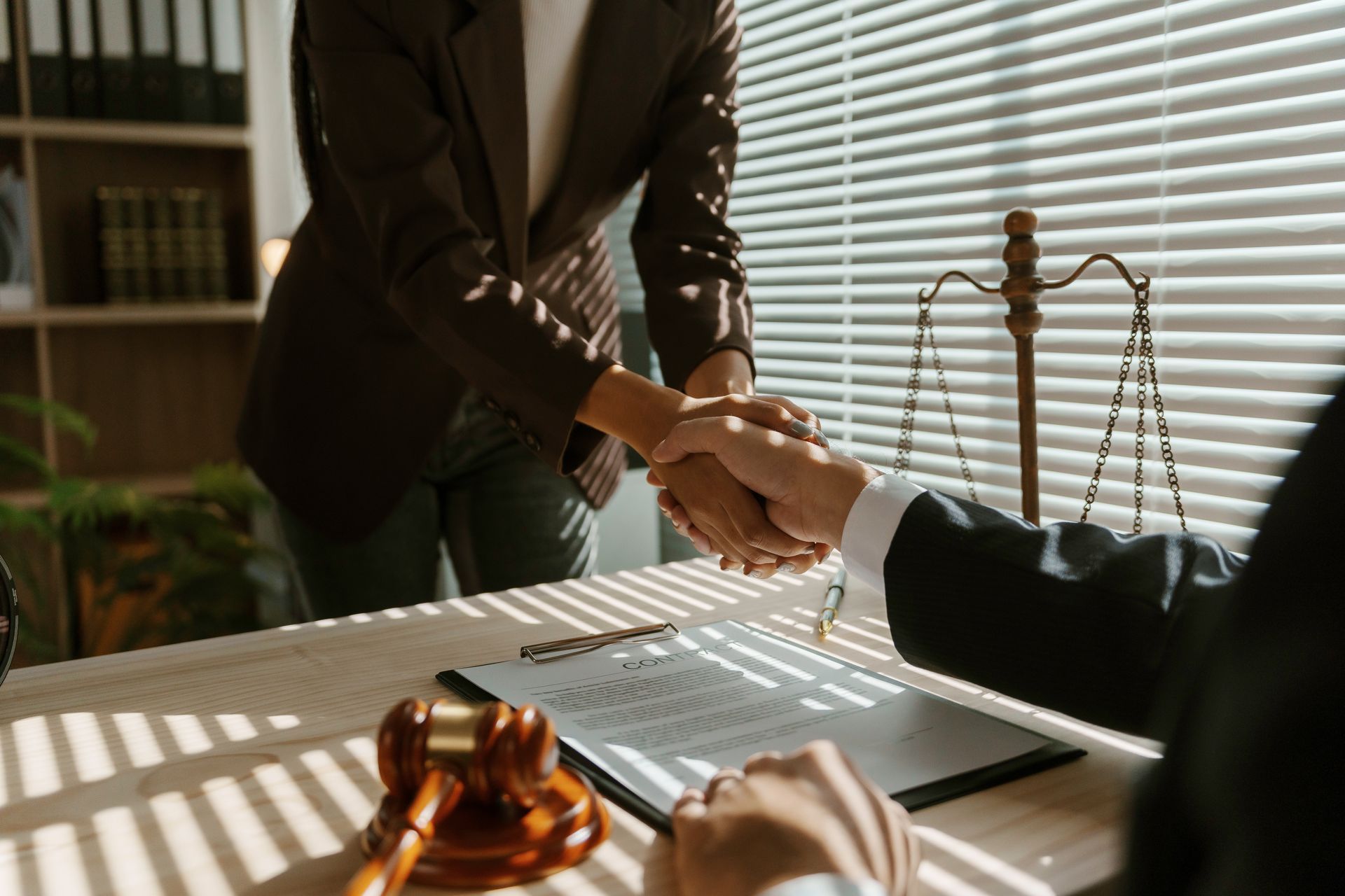 Two people shaking hands over a desk with a gavel, document, and scales of justice.