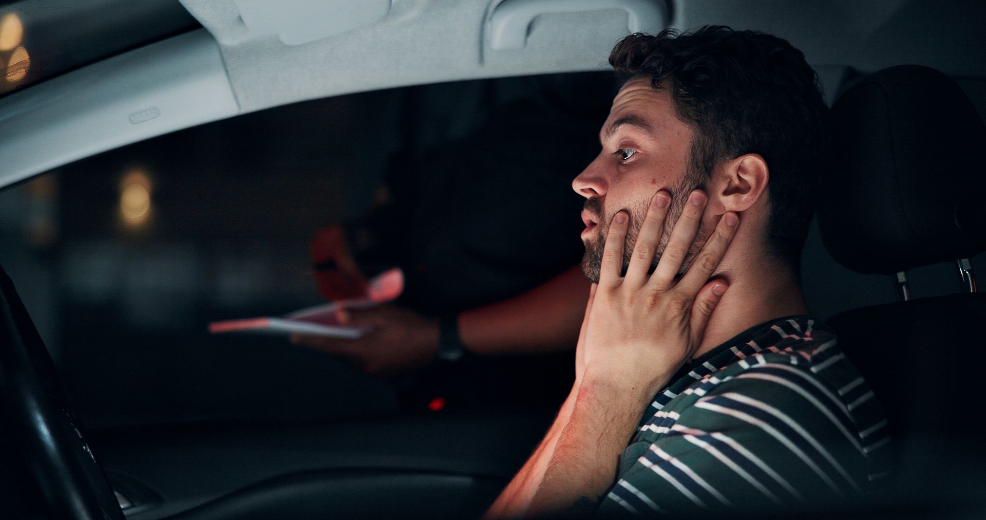 A man in a striped shirt sits in a car at night, looking distressed as an officer hands him a ticket through the window.
