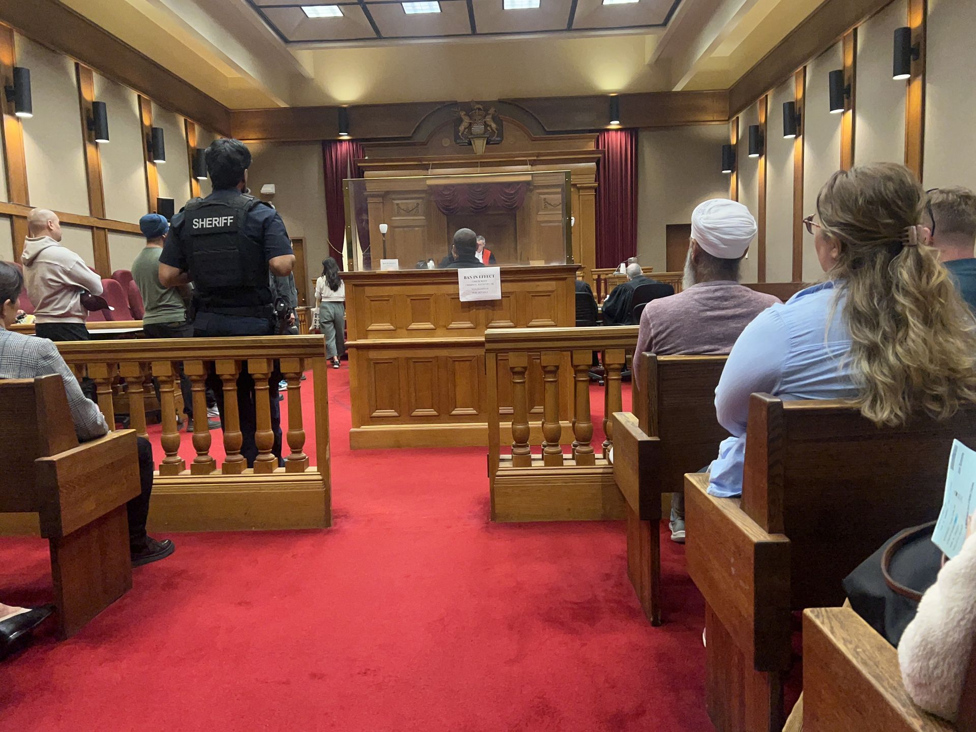 A courtroom with people seated, red carpet, wooden benches, and a judge's bench. A security guard stands.