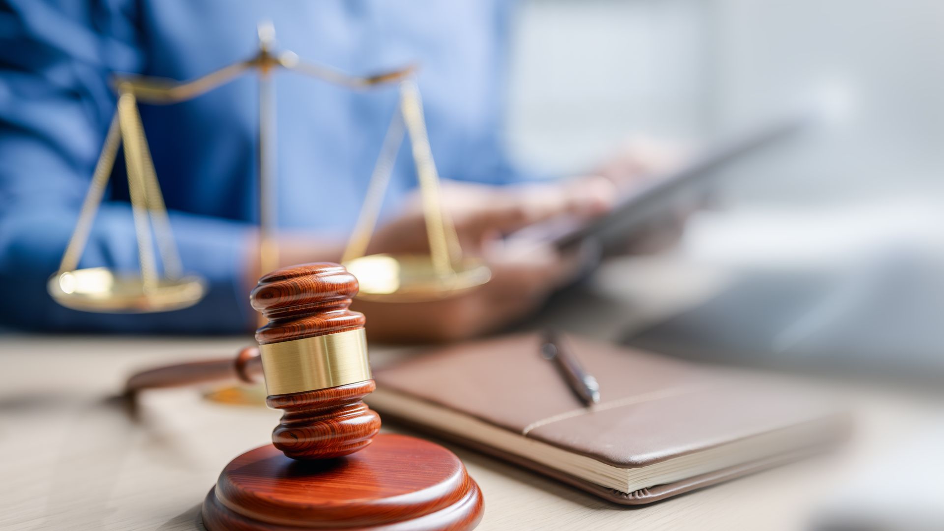 Gavel, scales of justice, notepad, and pen on a desk. Person using a tablet in background.