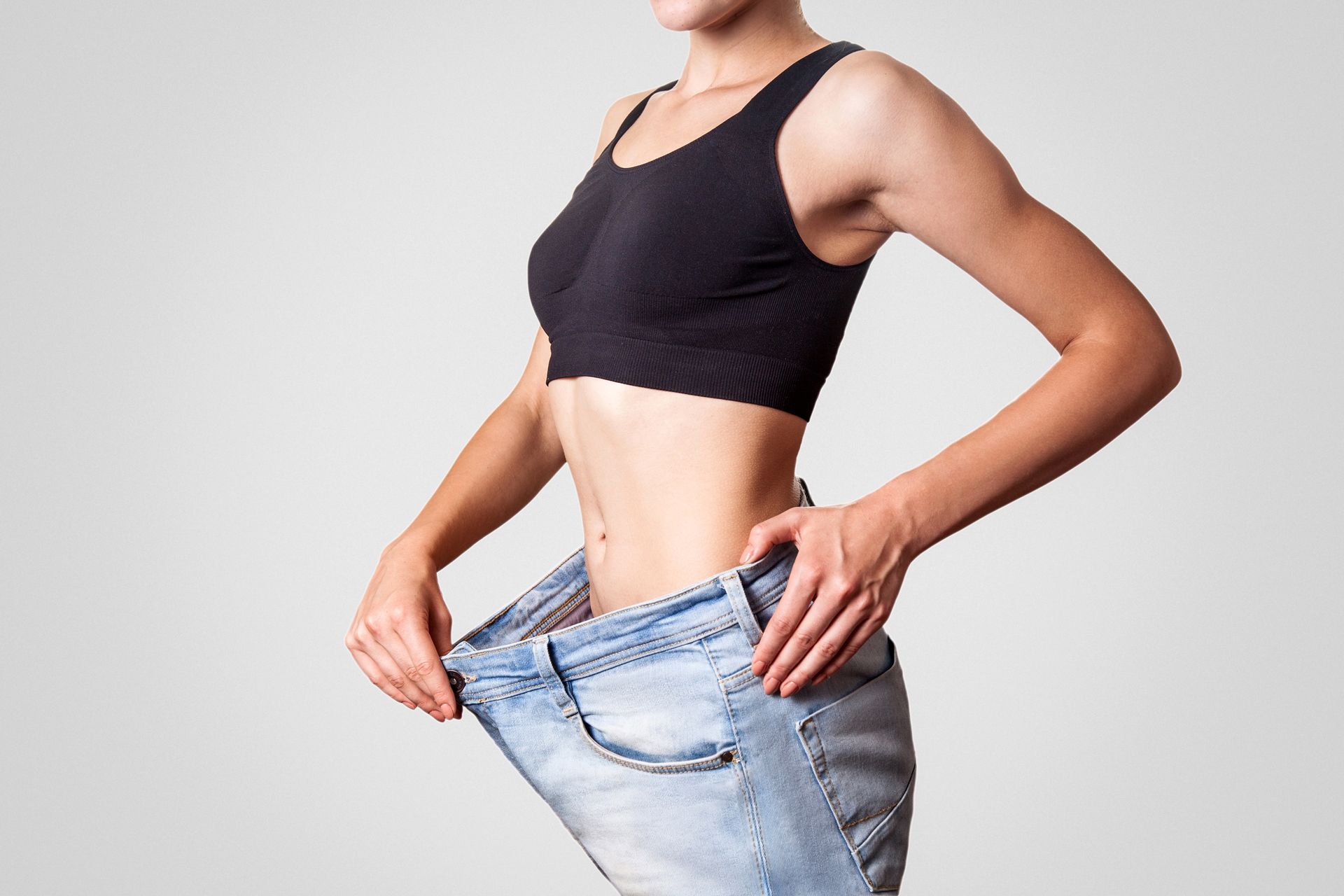 Woman in black sports bra pulling loose blue jeans around her waist