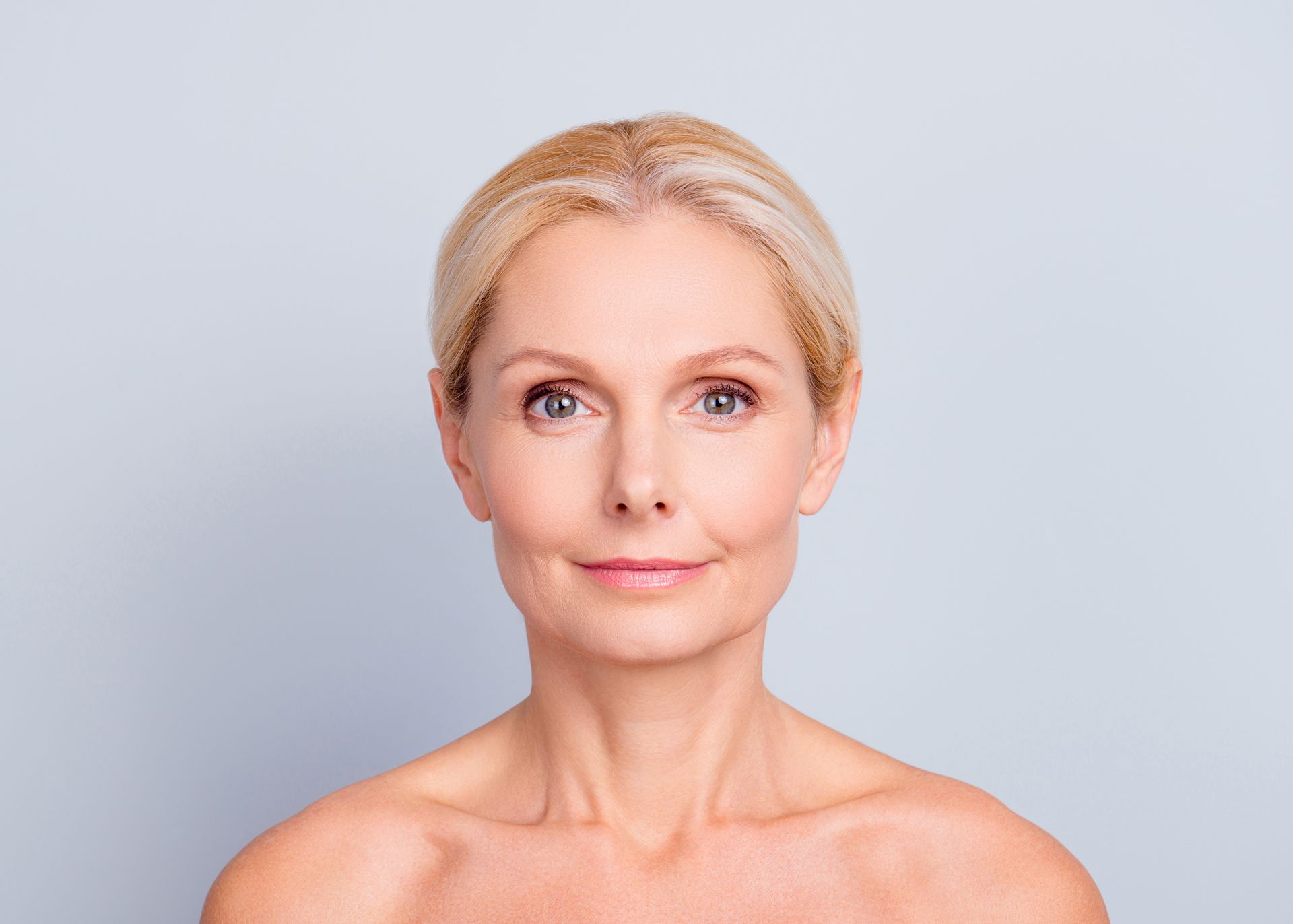 Blond woman with smooth skin, bare shoulders, and a slight smile against a light blue background.