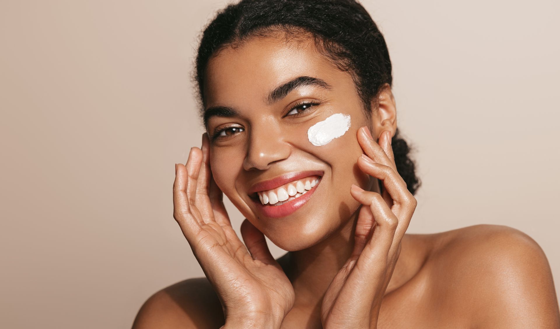 Woman applying cream to face, smiling, against a light beige background.