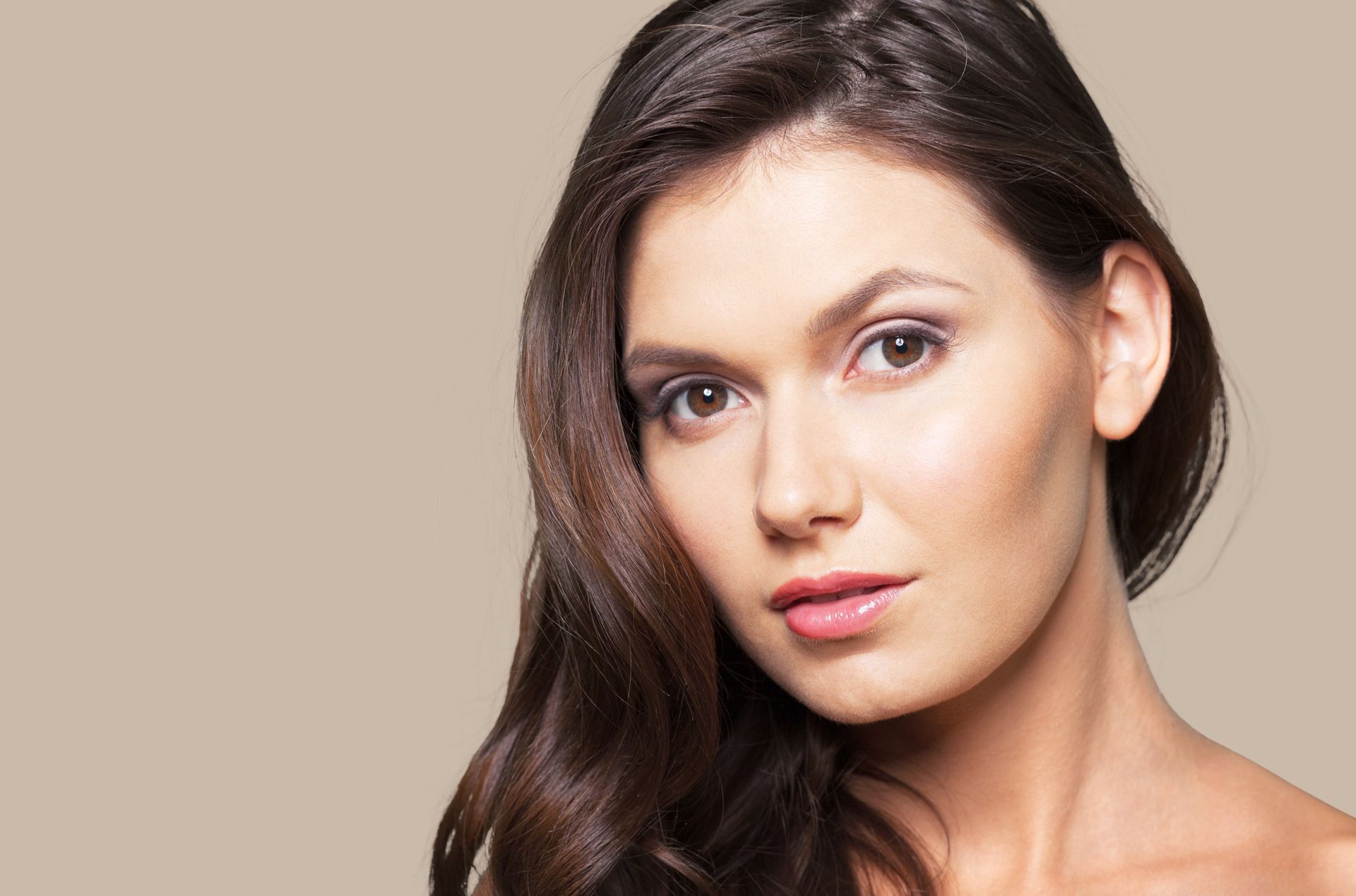 Woman with long, dark wavy hair and light skin looking towards the camera, against a neutral background.