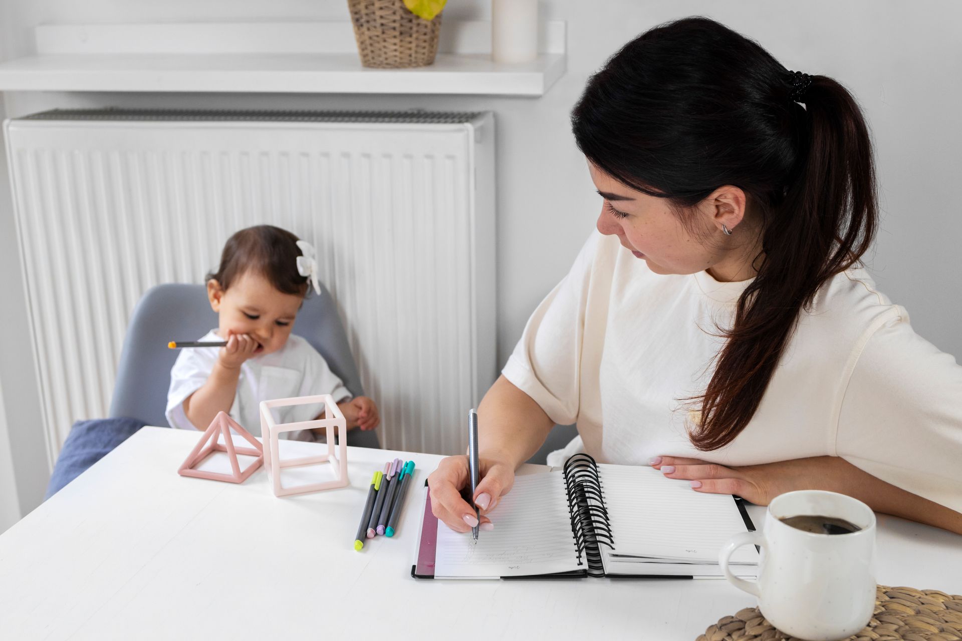 A woman is sitting at a table with a baby and writing in a notebook.