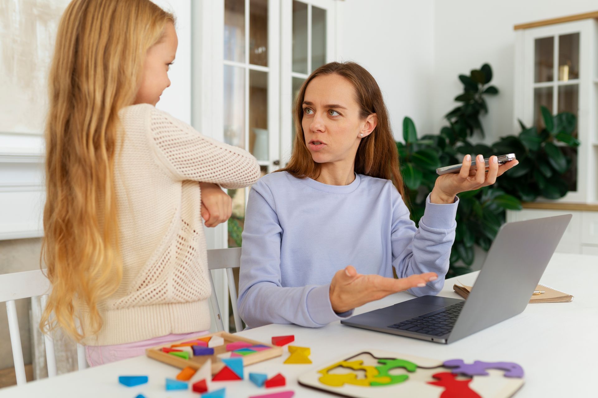 A woman is sitting at a table with a laptop and talking to a little girl.
