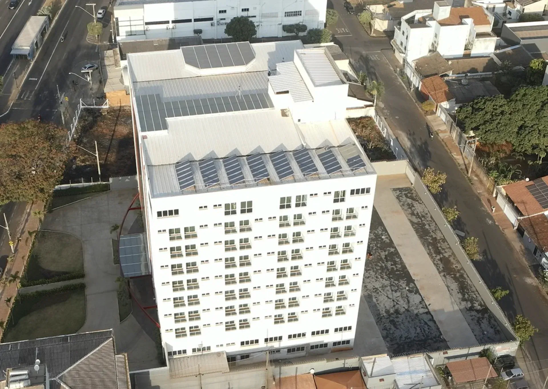Vista aérea de um prédio alto e branco com painéis solares no telhado; a rua e outros edifícios o rodeiam.