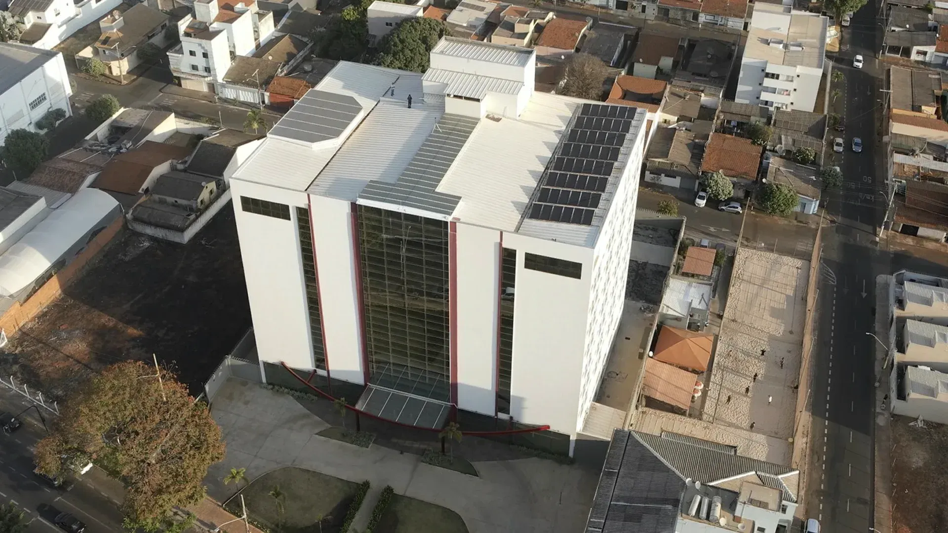 Vista aérea de um edifício moderno branco com painéis solares no telhado, rodeado por outros edifícios e uma estrada.