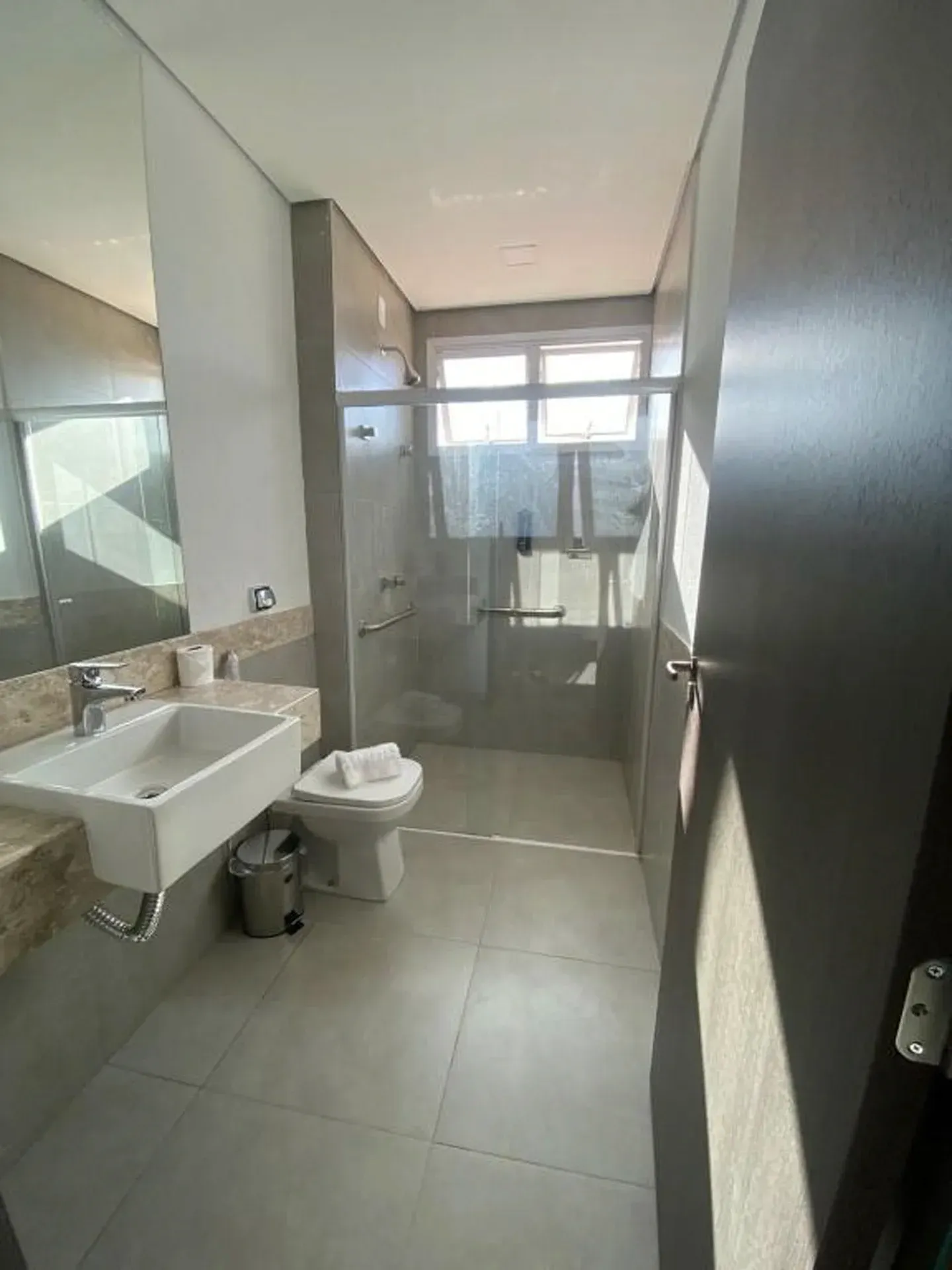 Banheiro com pia, vaso sanitário, box de vidro e janela. Paredes e piso em tom cinza claro.