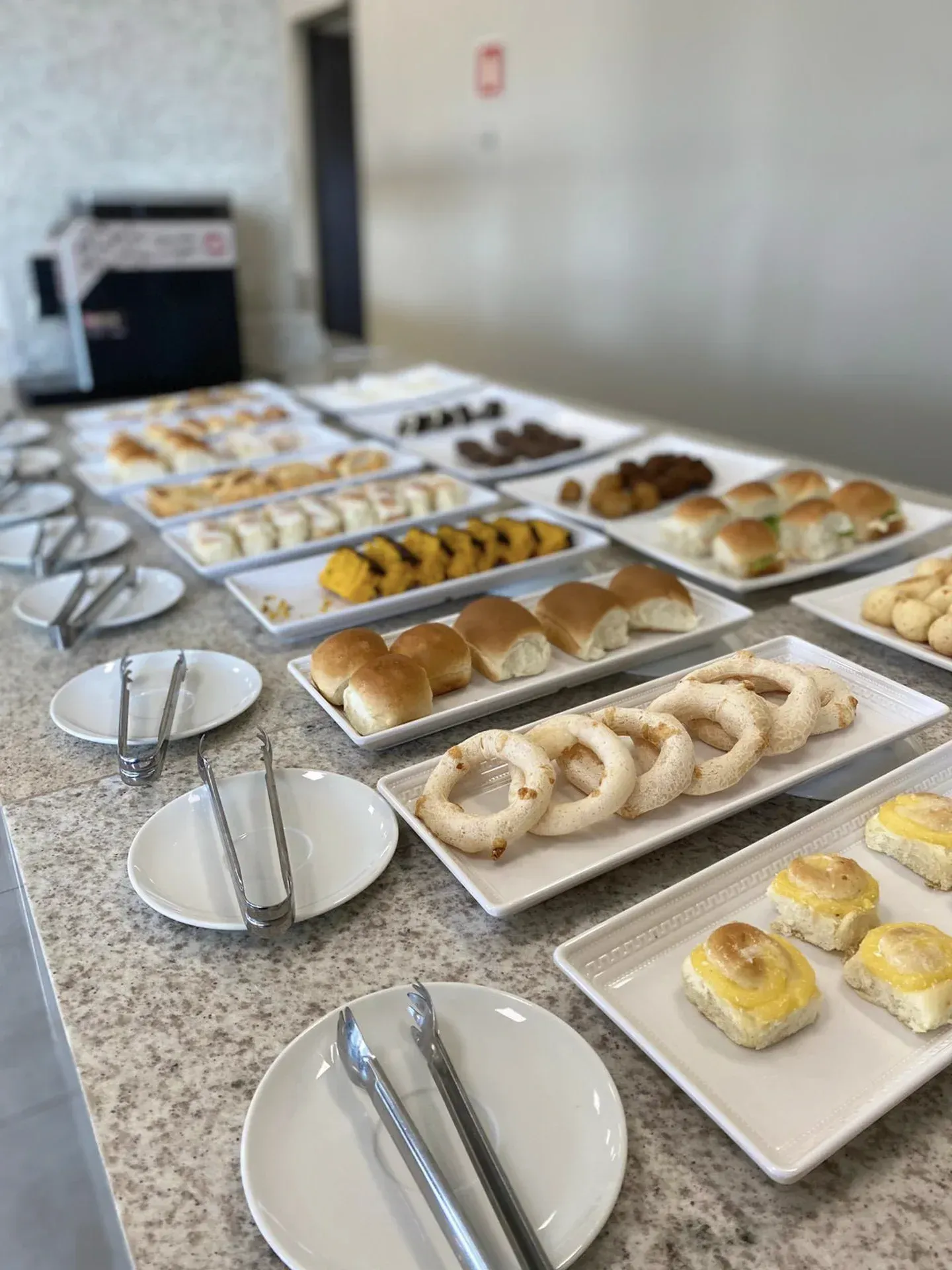 Mesa de buffet com diversos produtos de panificação: pãezinhos, doces e pães. Pratos brancos com pegadores estão disponíveis.