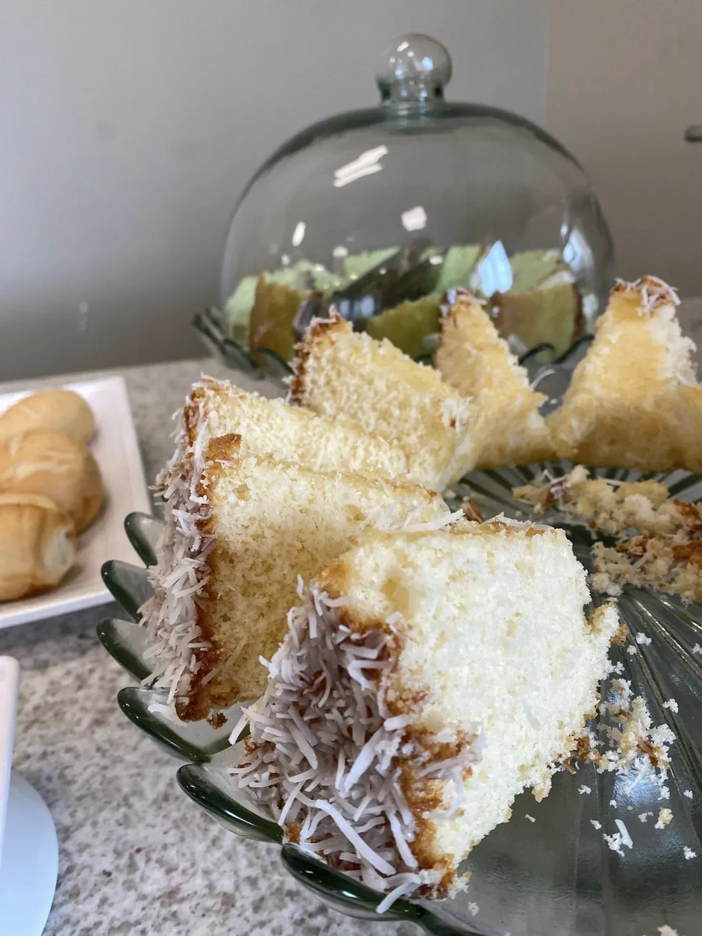 Bolo de coco fatiado em um prato de vidro, com outros alimentos ao fundo.