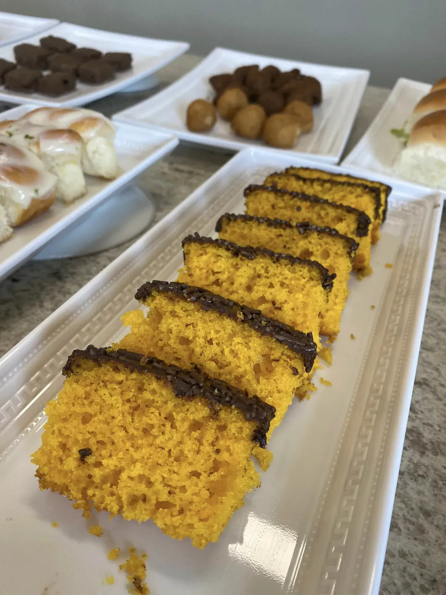 Fatias de bolo de laranja com cobertura de chocolate em um prato retangular branco; outras sobremesas ao fundo.