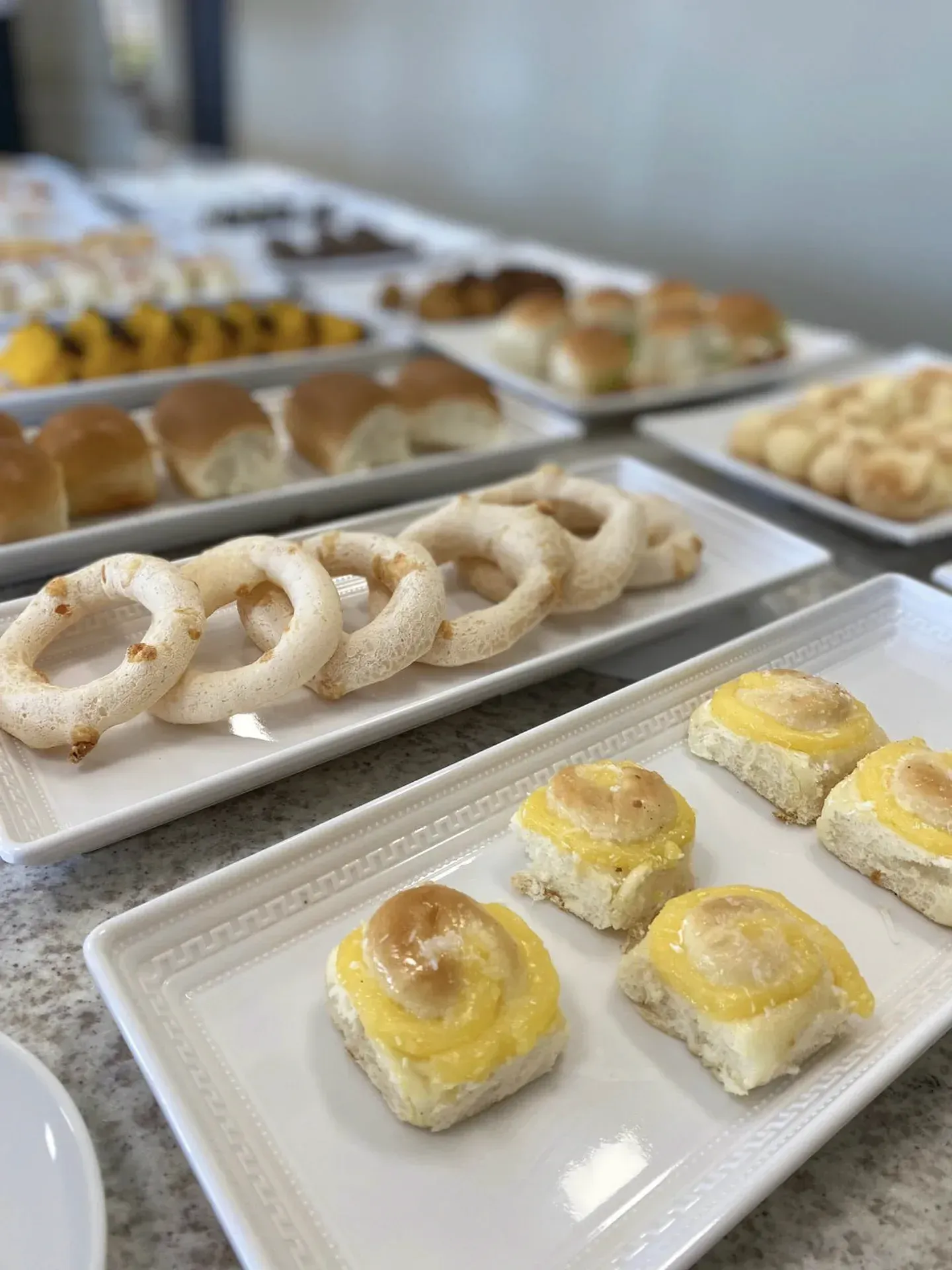 Bandejas com diversos produtos de panificação, incluindo pãezinhos, roscas e rolinhos de massa folhada.