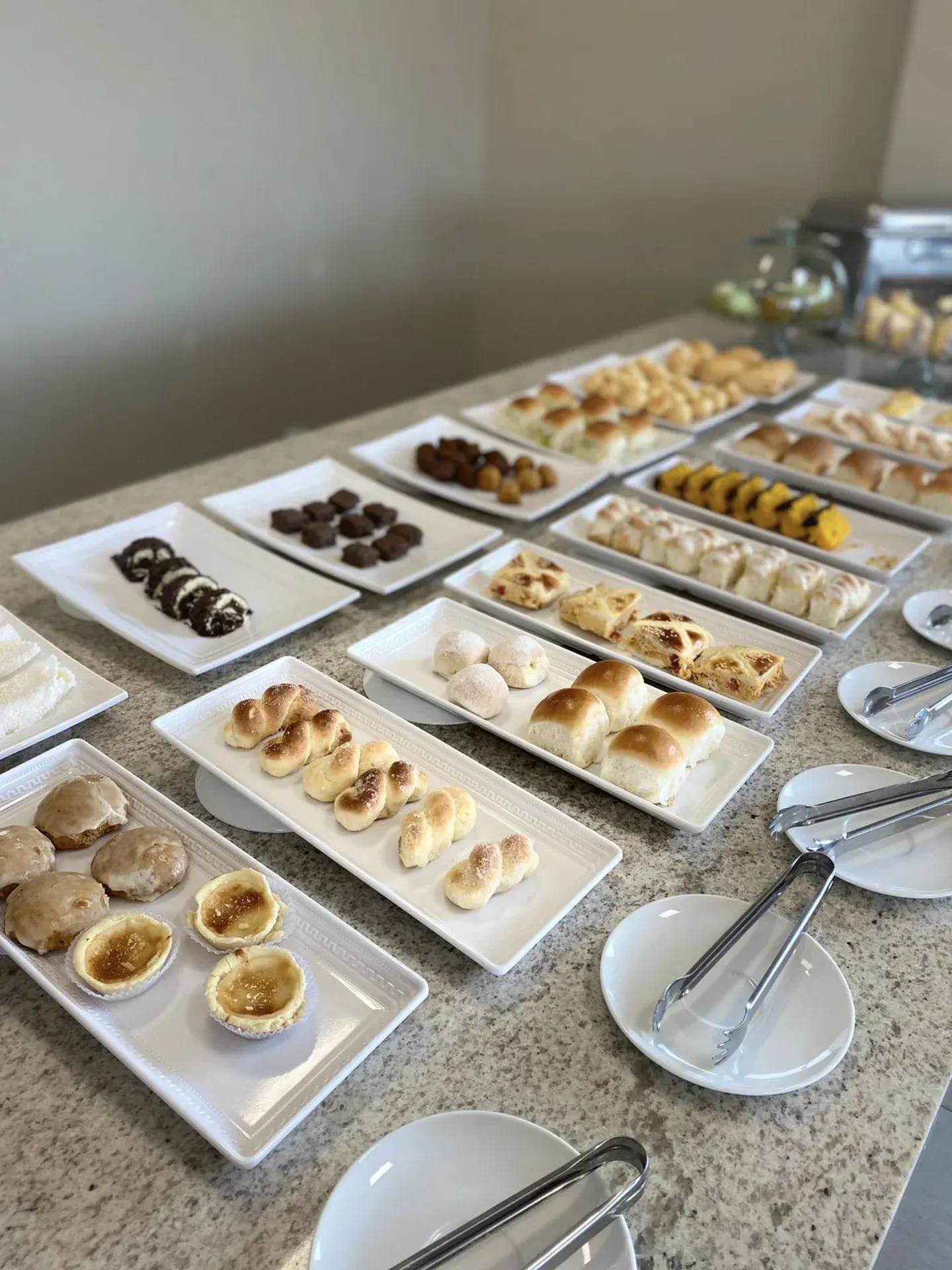 Mesa de bufê com diversos doces e pães em travessas retangulares brancas.
