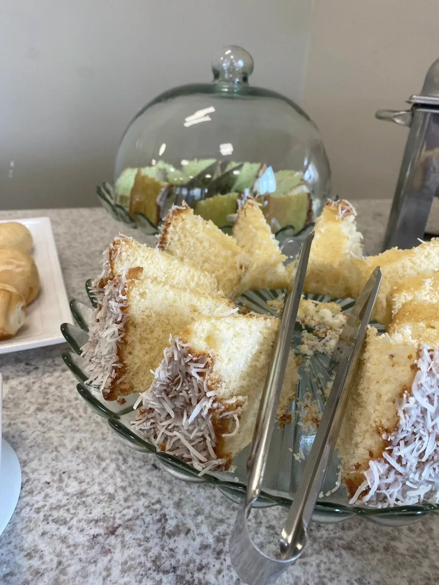 Fatias de bolo em um prato de vidro, com flocos de coco. Ao fundo, um prato coberto com mais bolo.