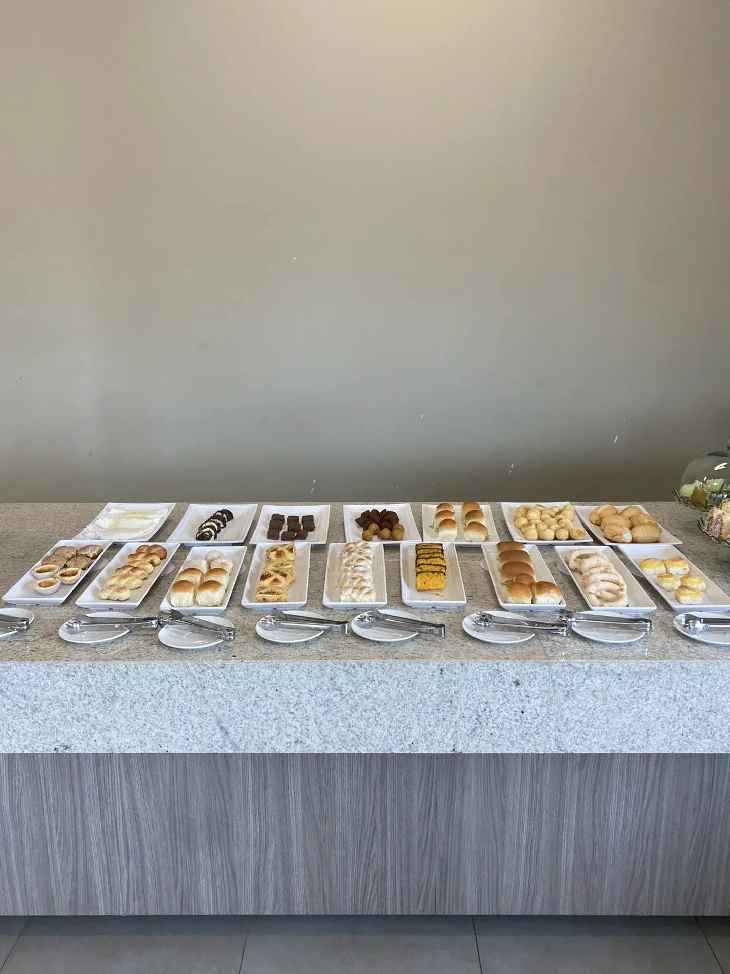 Uma mesa exibe diversos doces, dispostos em travessas com utensílios para servir, contra uma parede de cor neutra.