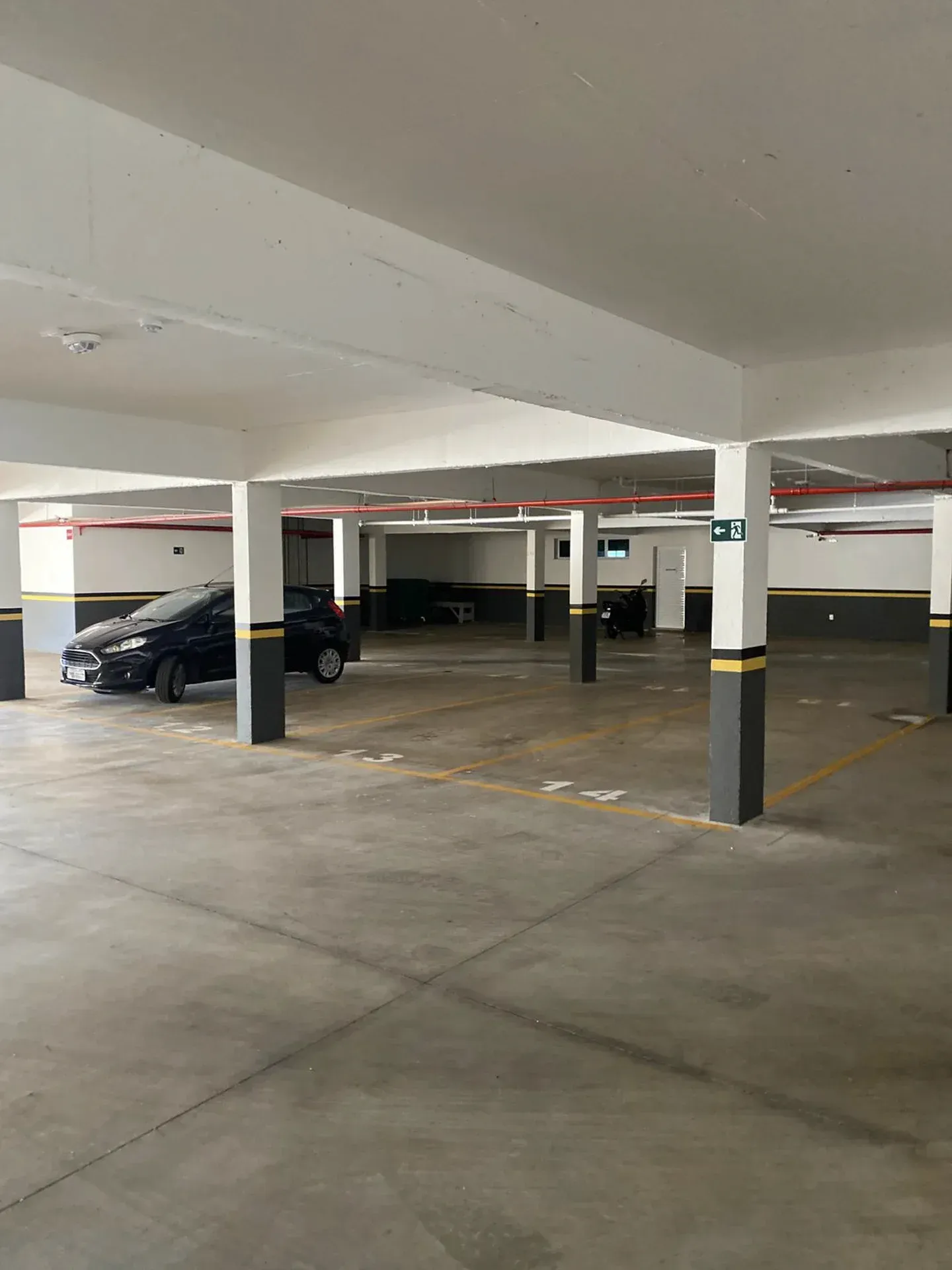 Garagem coberta; piso de concreto com carros estacionados, pilares de sustentação e iluminação no teto.