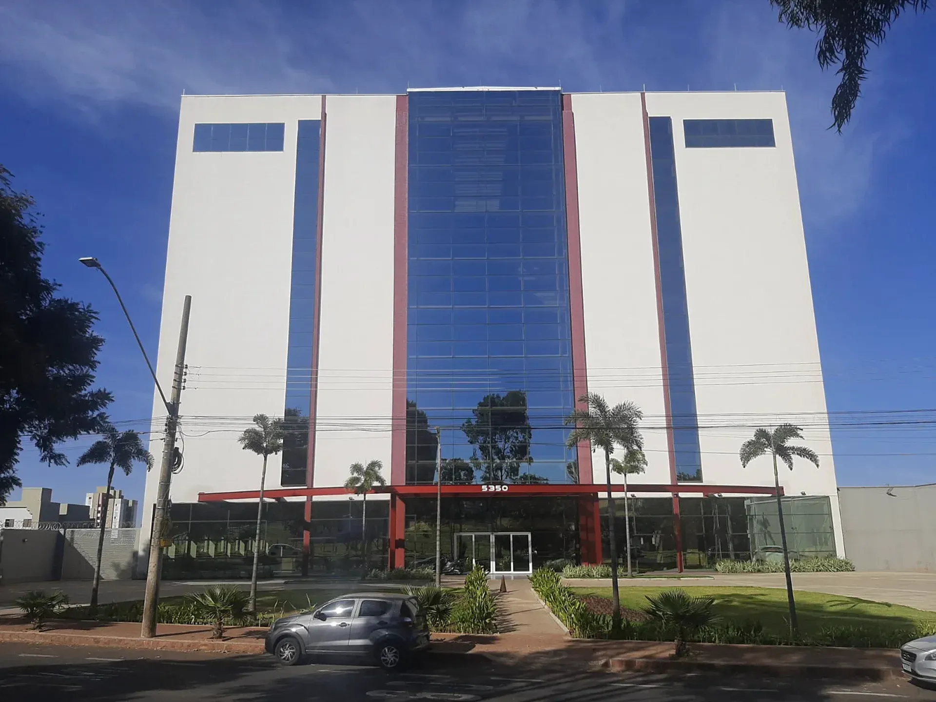Prédio de escritórios moderno com fachada de vidro e detalhes em vermelho, contra um céu azul.