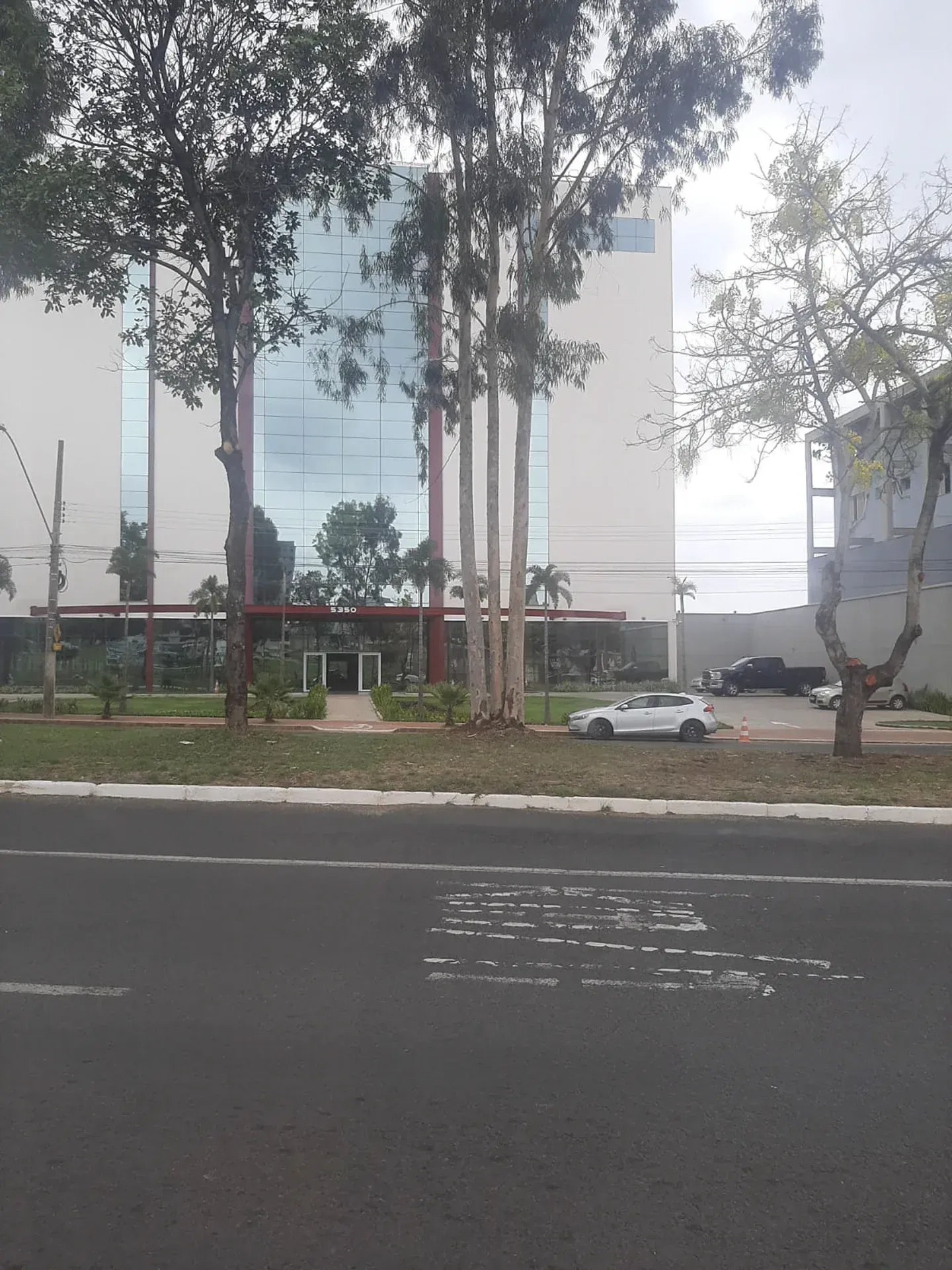 Prédio moderno com fachada de vidro e árvores na frente. Carro branco estacionado nas proximidades.