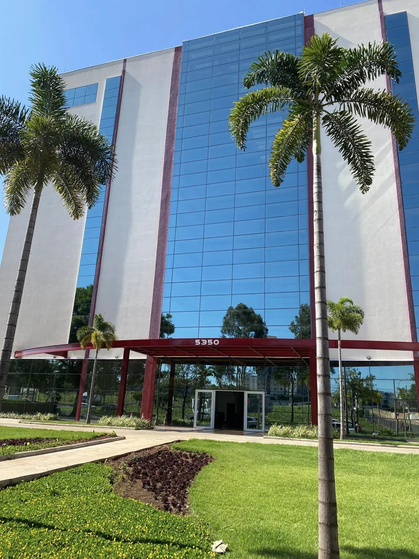 Prédio moderno com fachada de vidro azul, detalhes em vermelho e palmeiras. Entrada sinalizada com