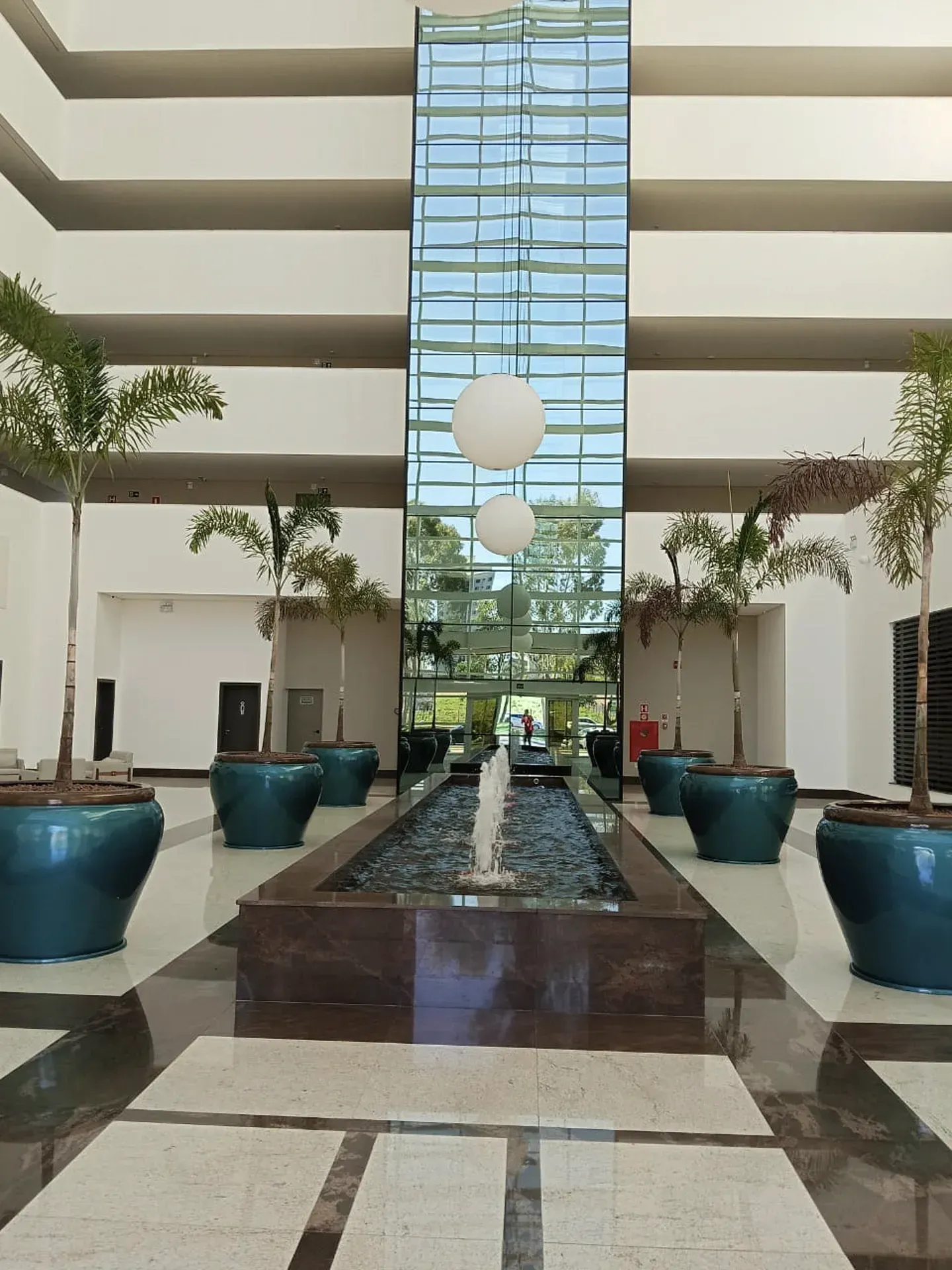 Hall de entrada com teto em níveis, fonte, plantas em vasos, poço de elevador envidraçado e luminárias redondas suspensas.