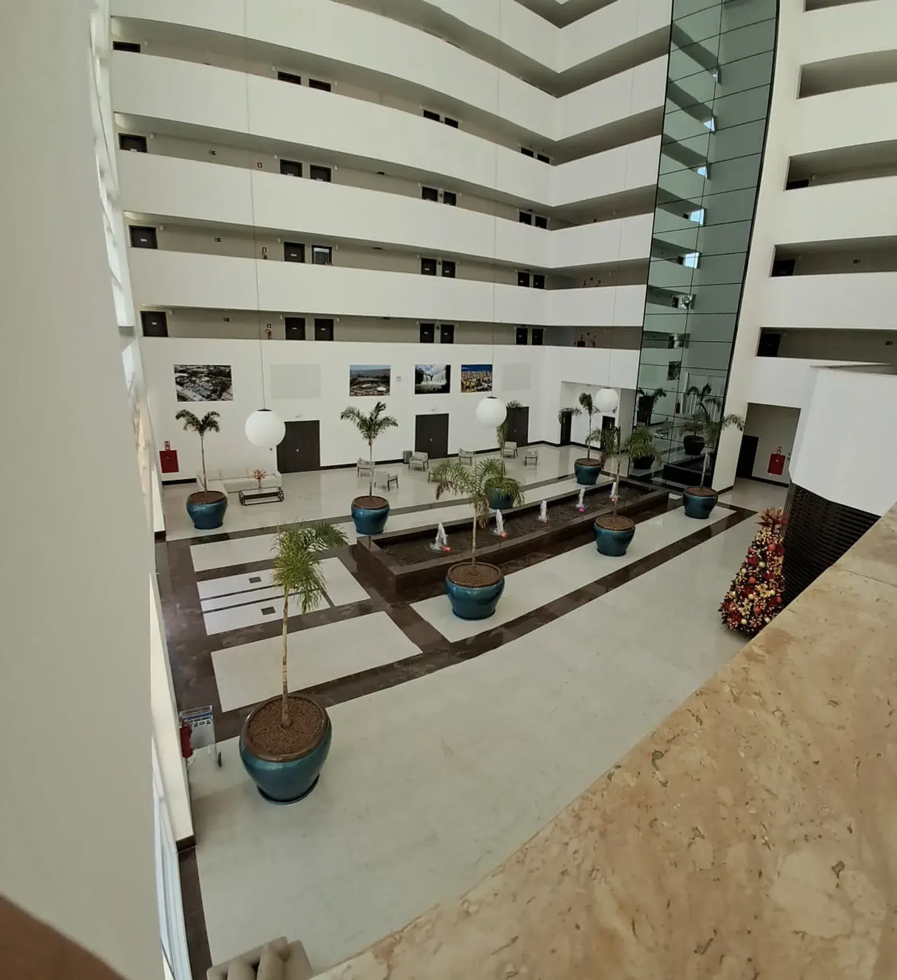 Vista interna do átrio de um edifício de vários andares com paredes brancas, varandas e plantas.