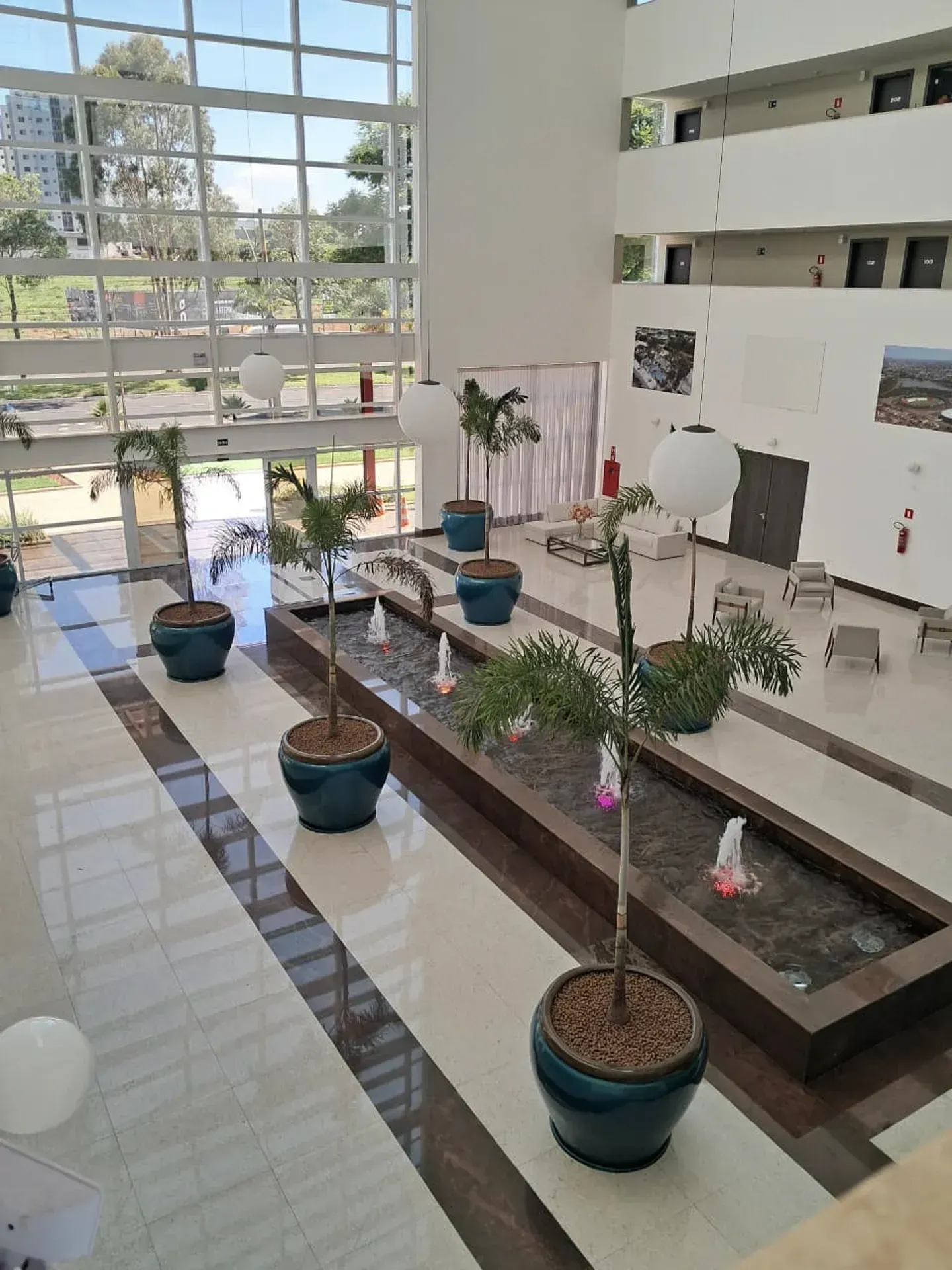 Hall de entrada com fonte e vasos de plantas; janelas altas e paredes brancas.