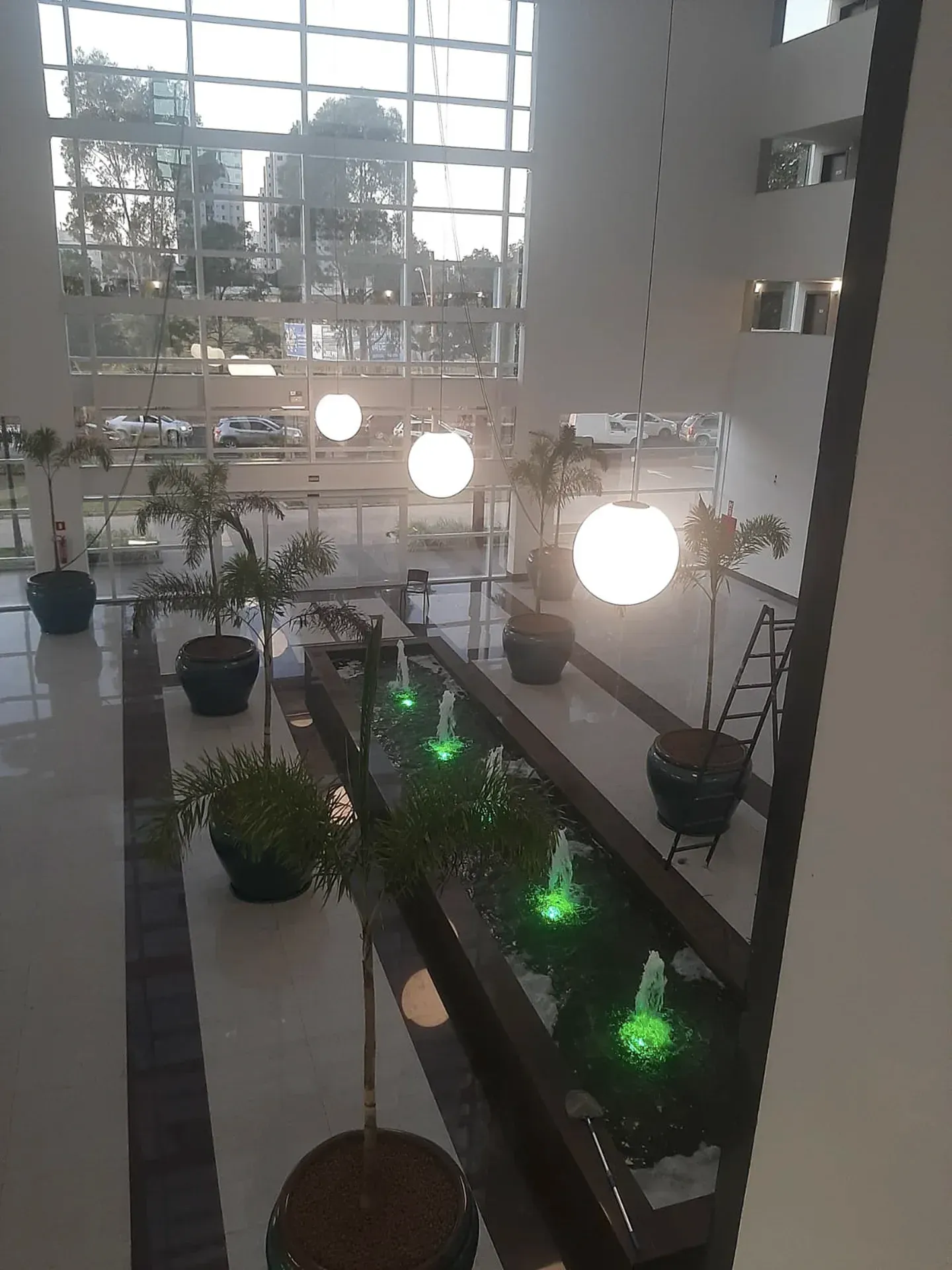 Vista interna do hall de entrada de um edifício moderno com fonte, plantas em vasos e luminárias globulares.