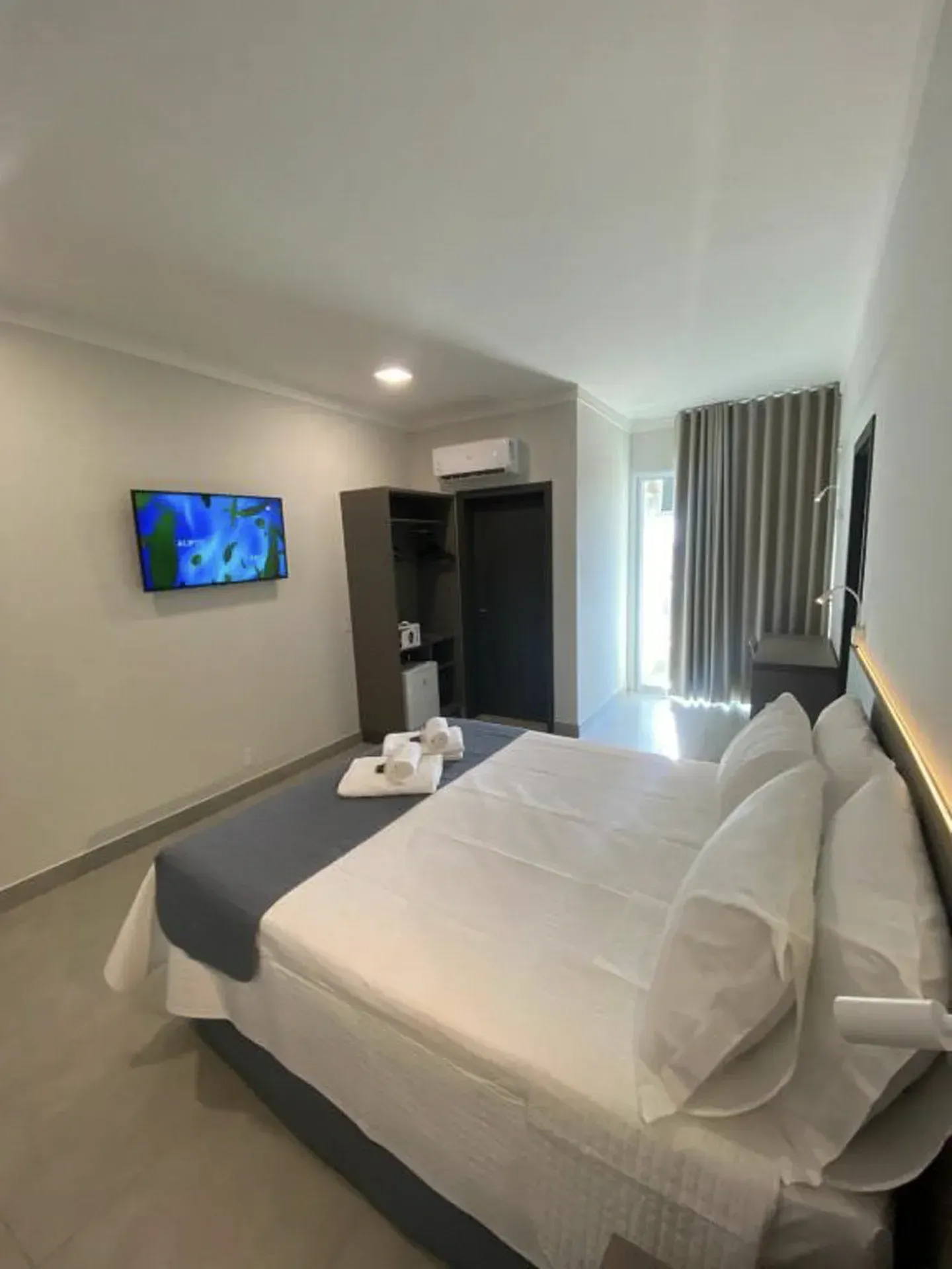 Quarto de hotel com cama, TV, armário e janela com cortinas.