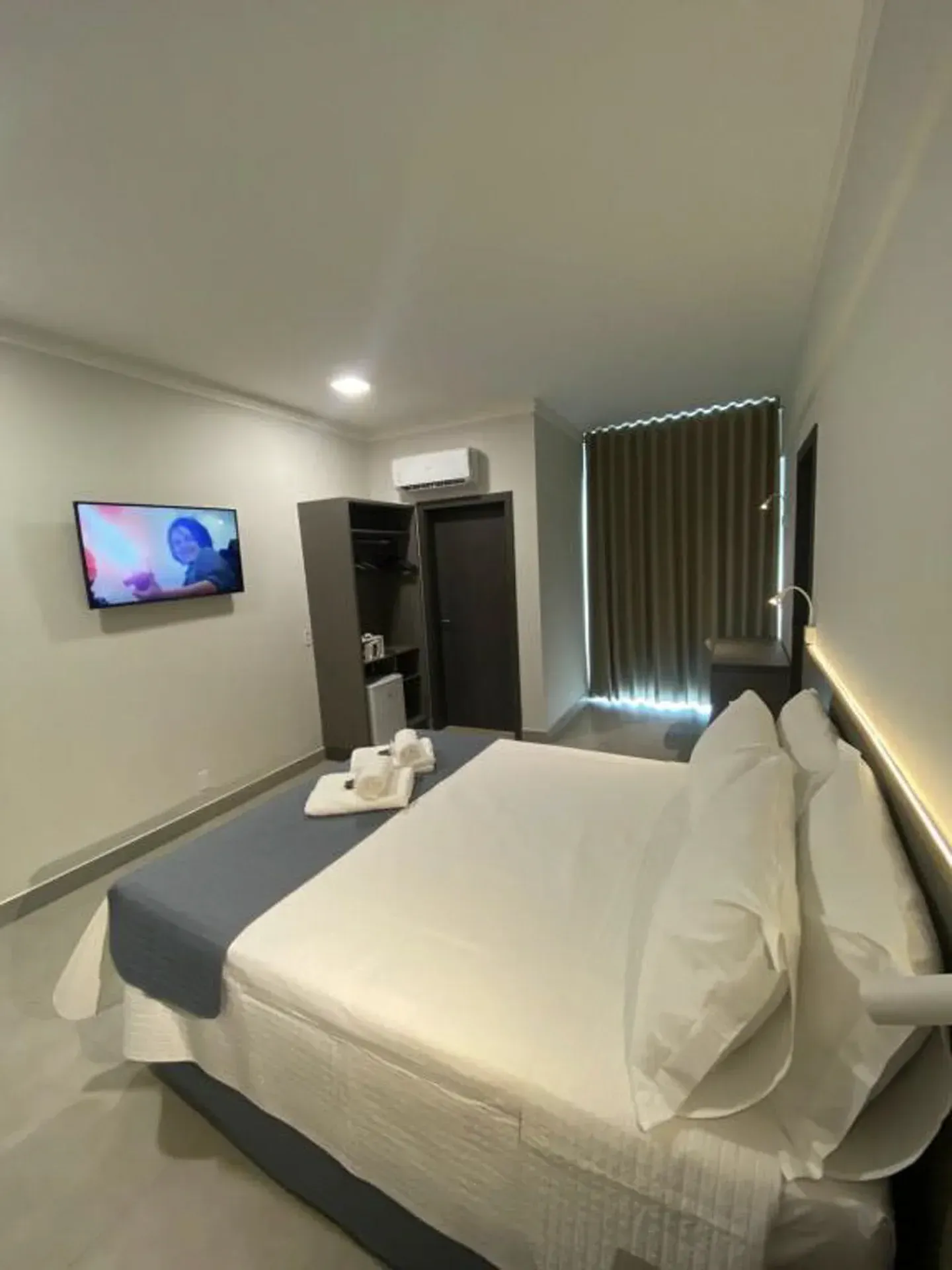 Quarto de hotel com cama, TV, armário e janela. Lençóis brancos, paredes cinza e cobertor azul.