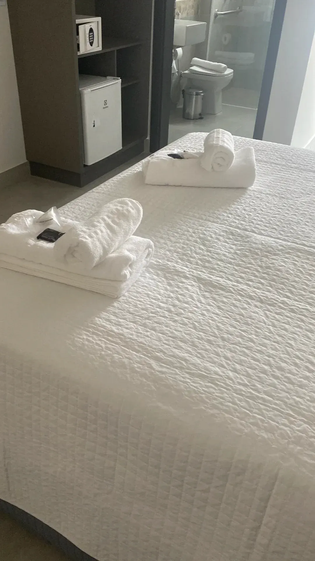 Cama branca com toalhas dobradas; frigobar e banheiro ao fundo.