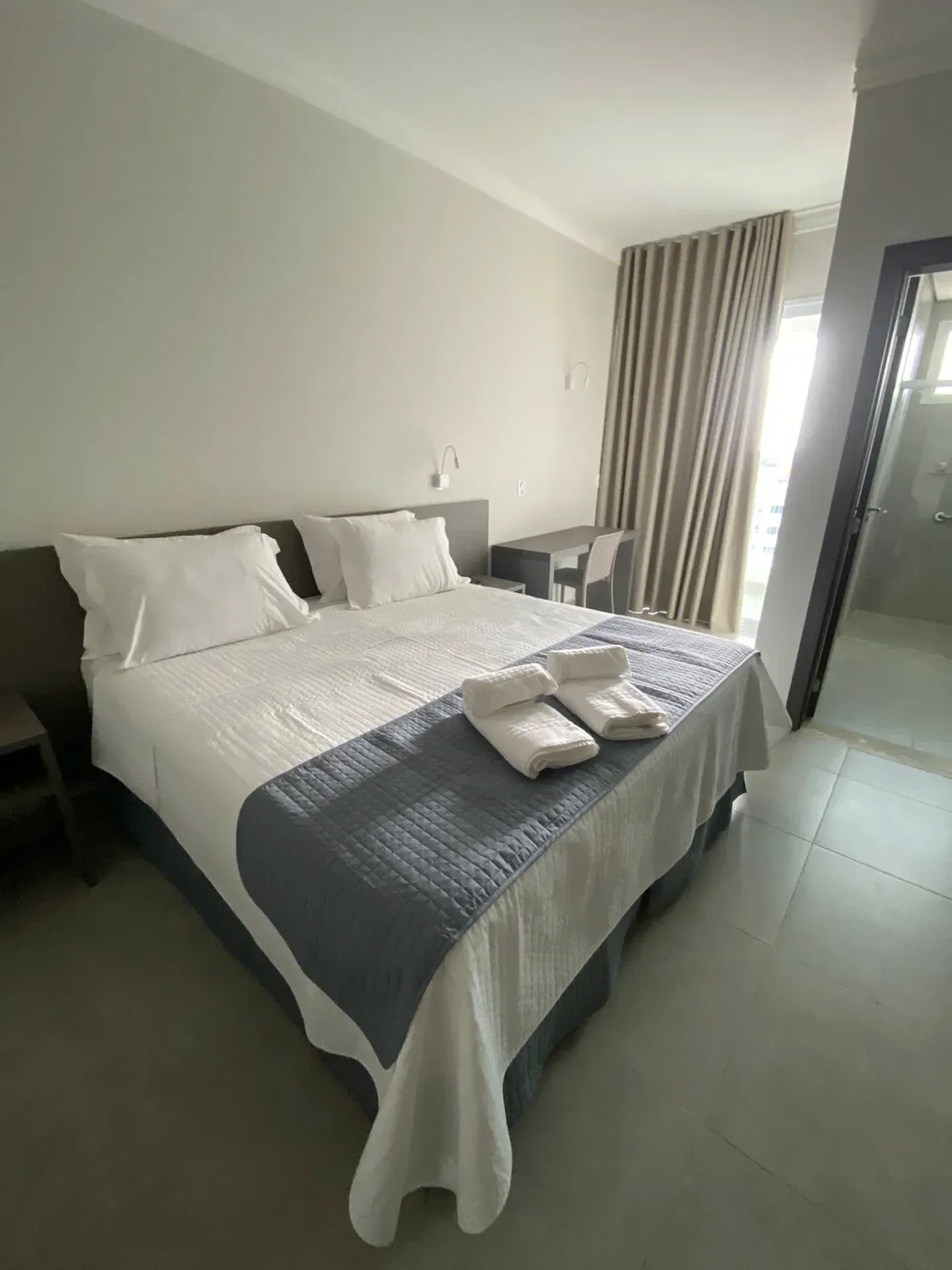 Quarto com cama grande, lençóis brancos e tapete azul. Paredes, cortinas e escrivaninha em tom cinza claro. Porta visível.