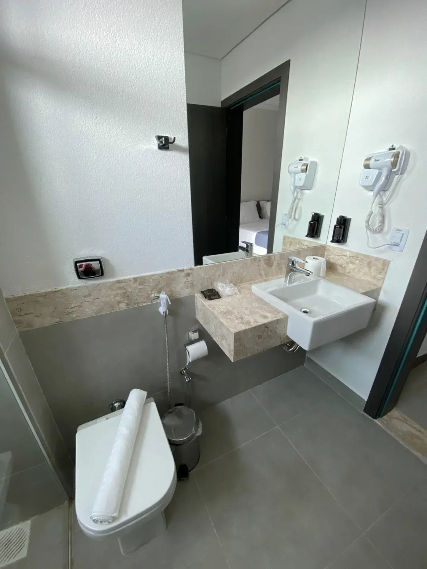 Banheiro com vaso sanitário, pia, espelho e secador de cabelo. Tons de cinza e bege.