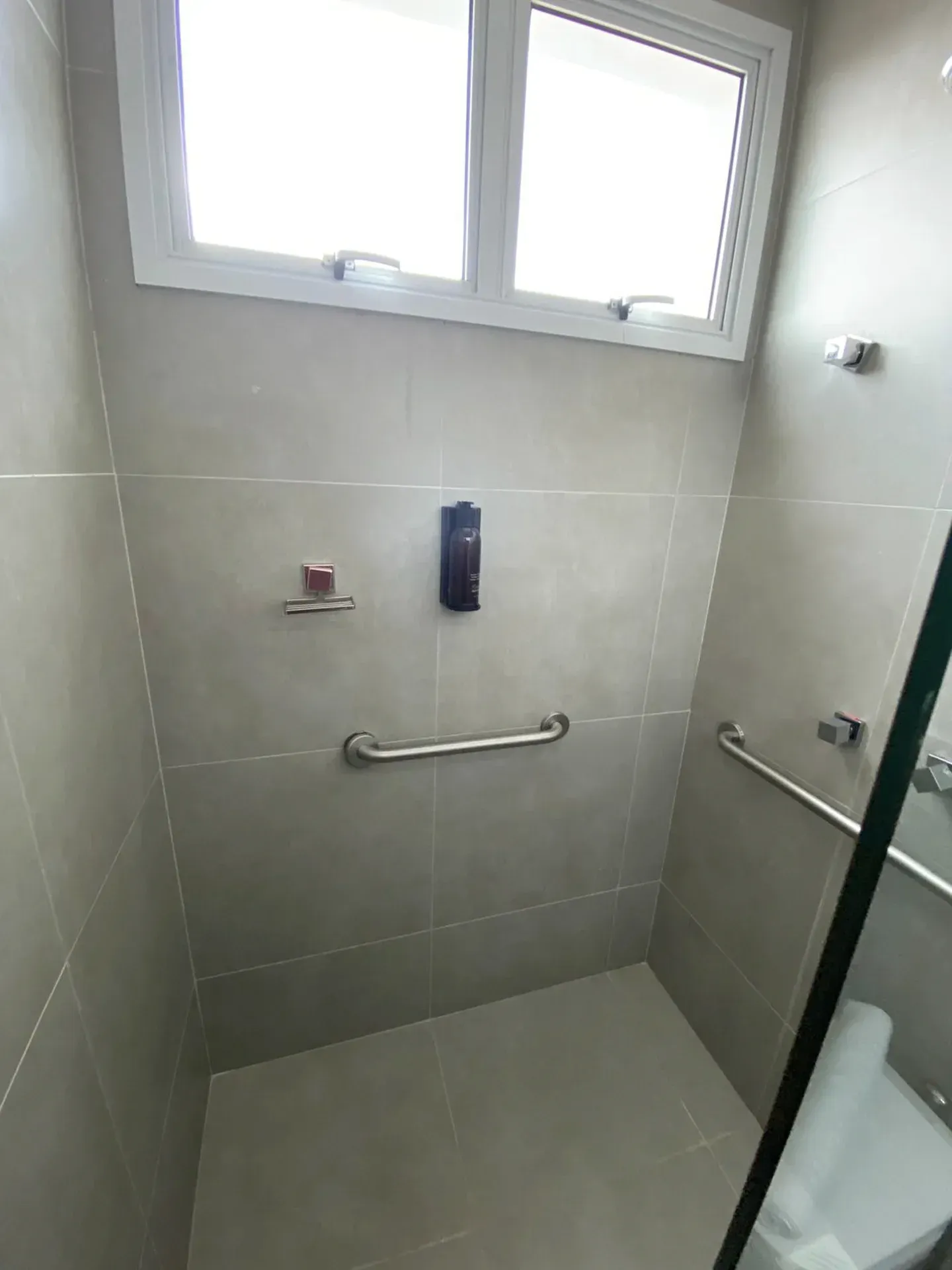 Chuveiro acessível com barras de apoio e dispensador de sabonete, azulejos cinza, janela pequena.
