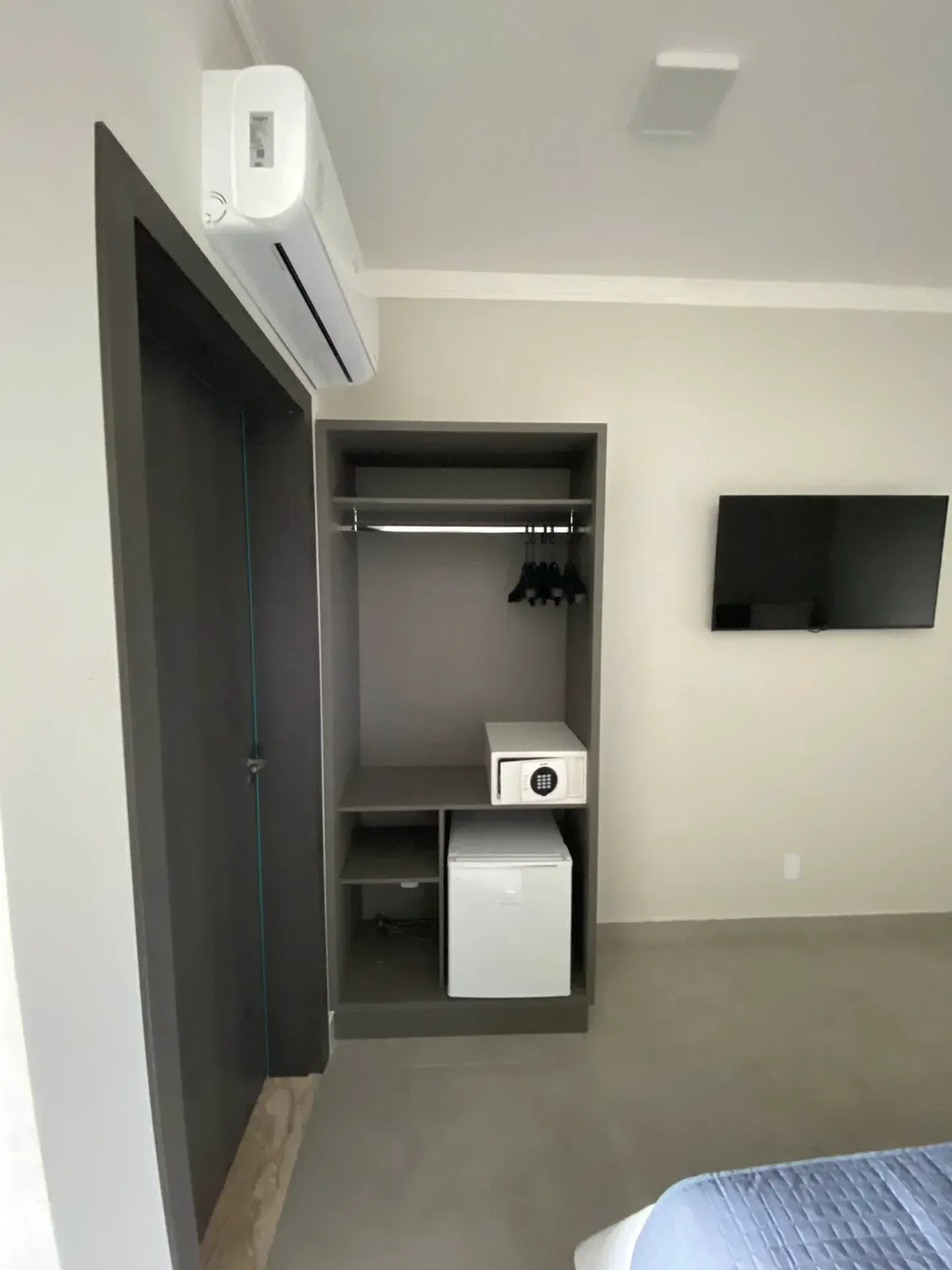 Armário embutido com varão para pendurar roupas, cofre e frigobar em um quarto com ar condicionado e TV fixada na parede.