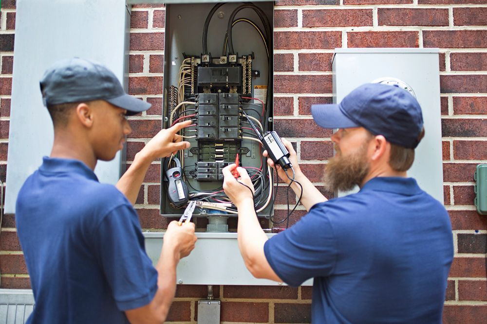 Two men are working on an electrical box on a brick wall.