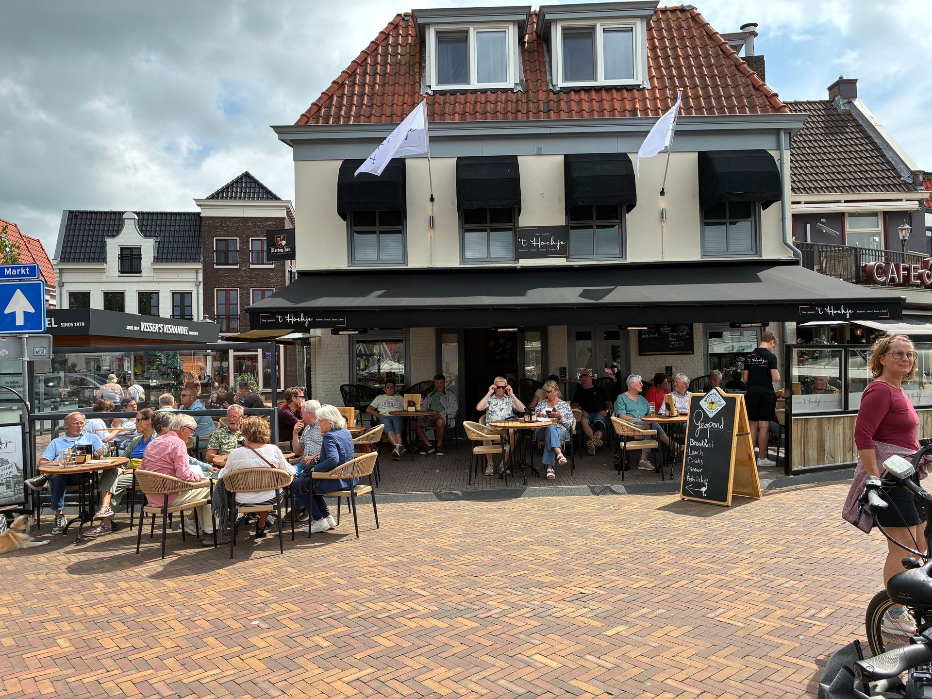 Mensen op een terrasje op een bakstenen plein. Gebouw met zwarte luifels en vlaggen. Zonnige dag.