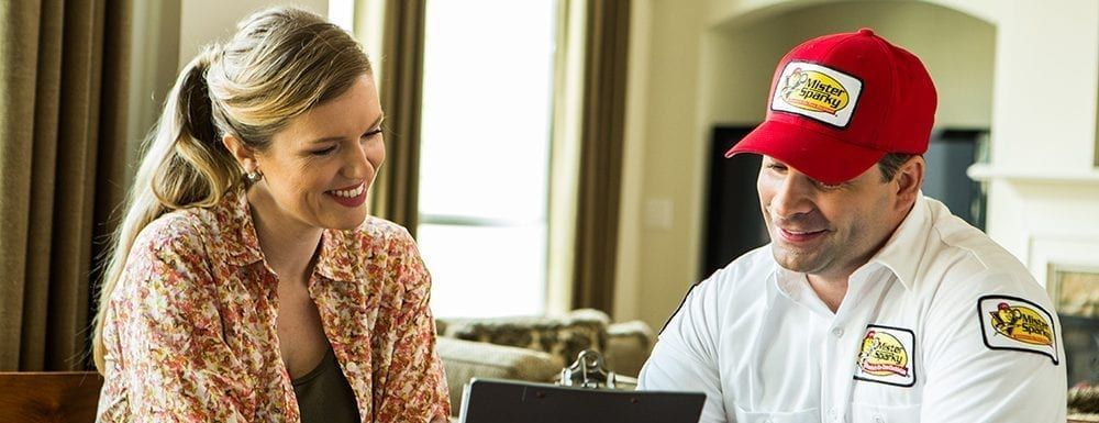 A man in a red Mister Sparky hat is talking to a woman in a living room.